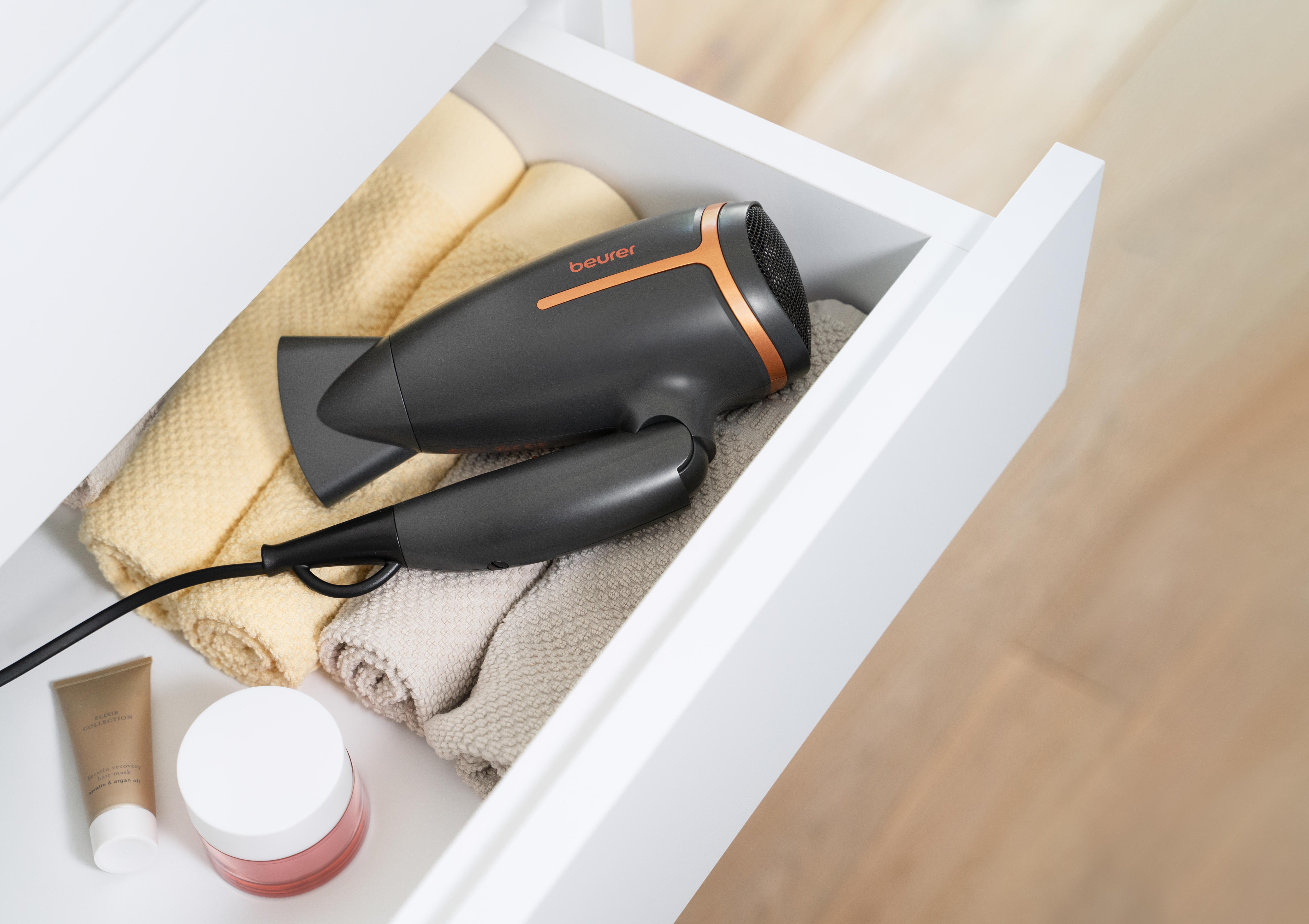 Ein Haartrockner, Handtücher und Hautpflegeprodukte liegen in einer weißen Schublade. Der Fön ist schwarz-goldfarben.