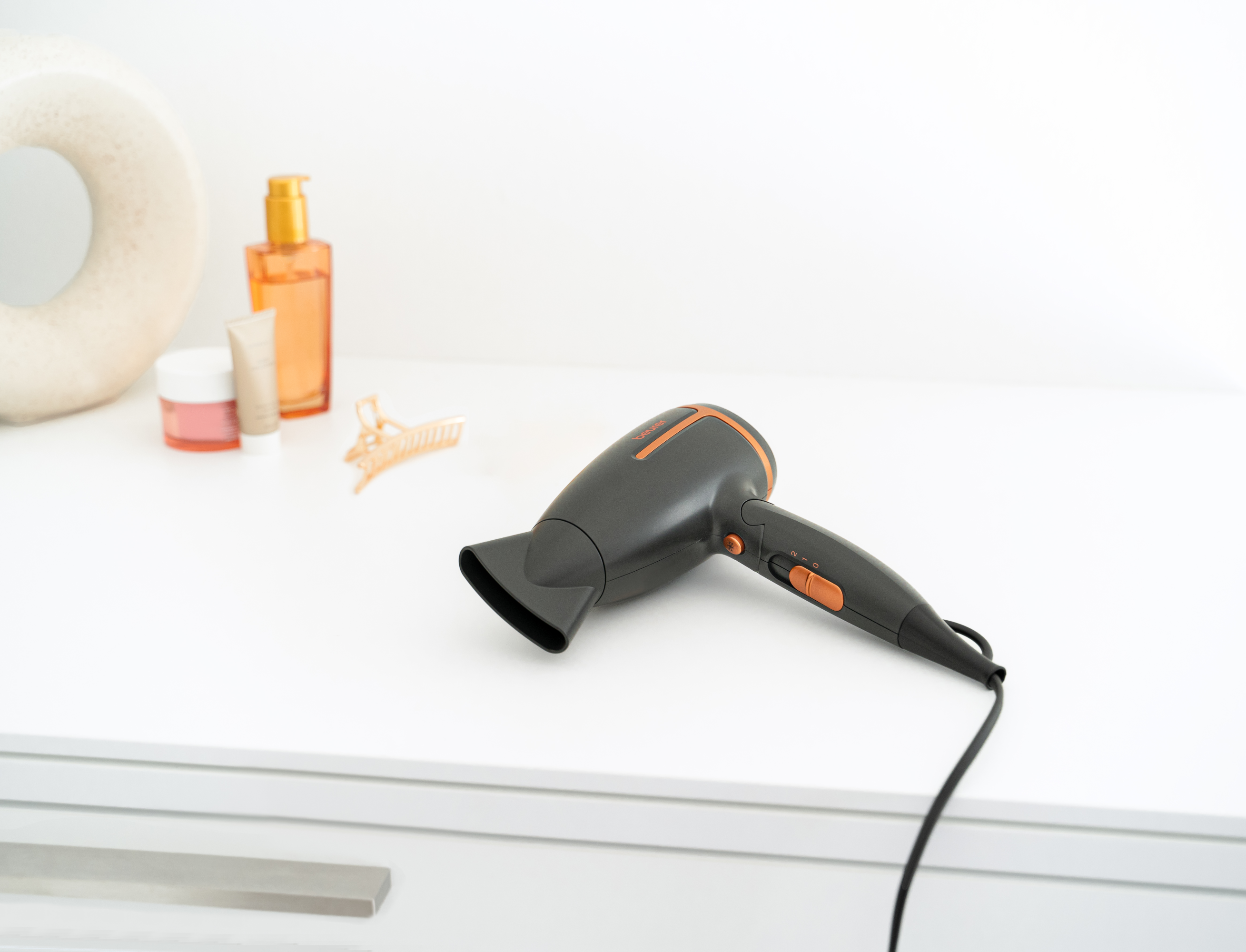 Ein schwarzer und orangefarbener Haartrockner auf einer weißen Oberfläche mit Beauty-Produkten im Hintergrund.