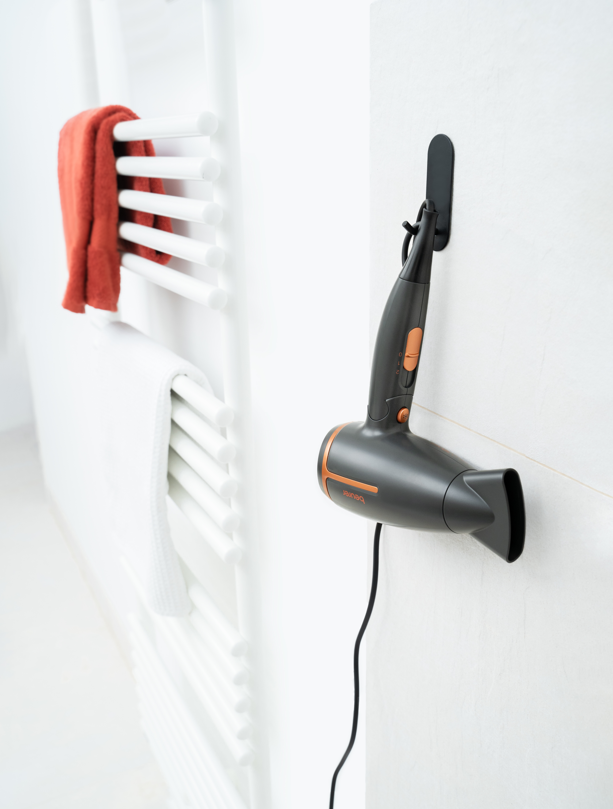 Ein Föhn hängt an einer weißen Wand neben einem Handtuchhalter mit einem roten Handtuch.