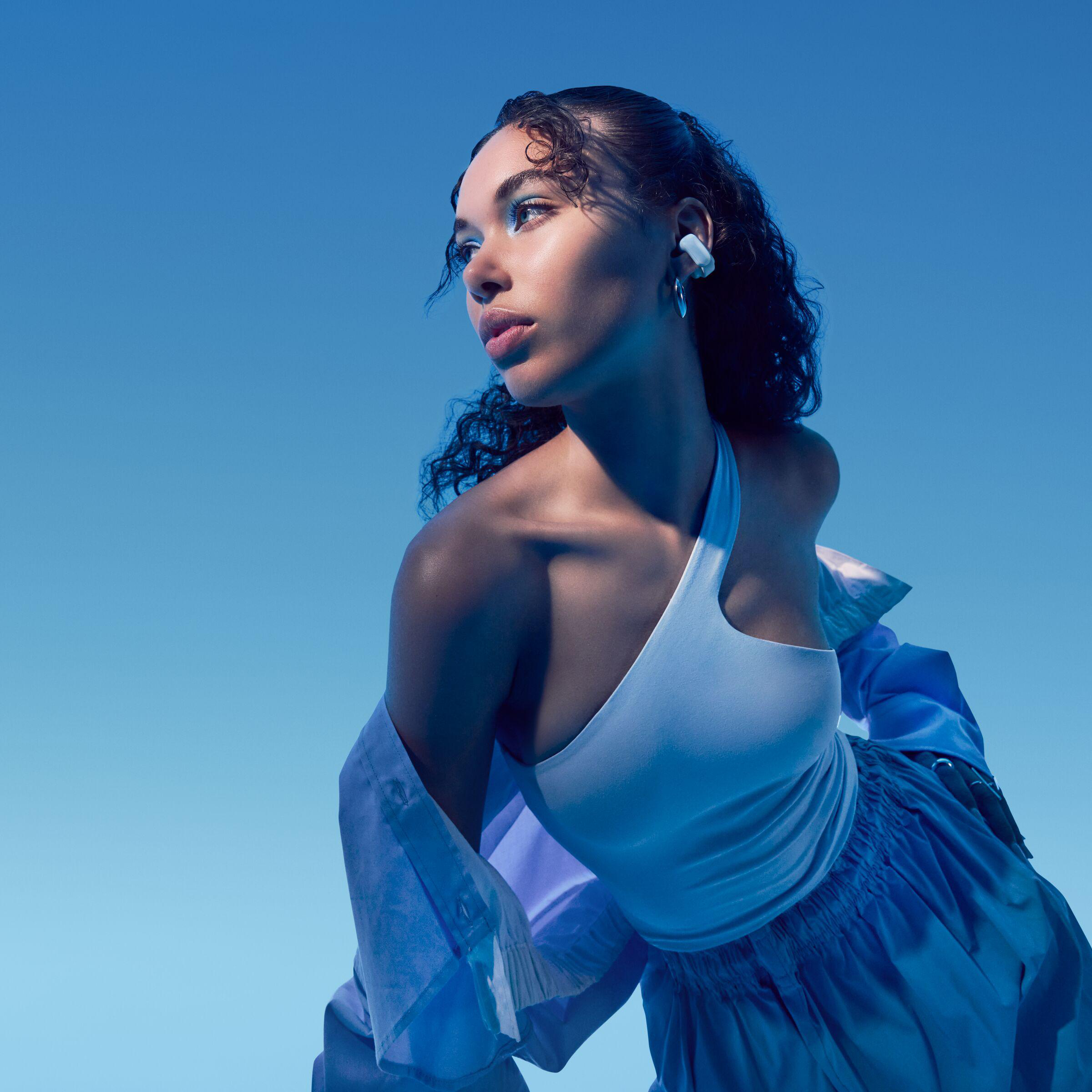 Eine Frau mit blauem Lidschatten und Ohrhörern, trägt ein weißes Oberteil und ein blaues Hemd, blickt weg. Der Hintergrund ist blau.