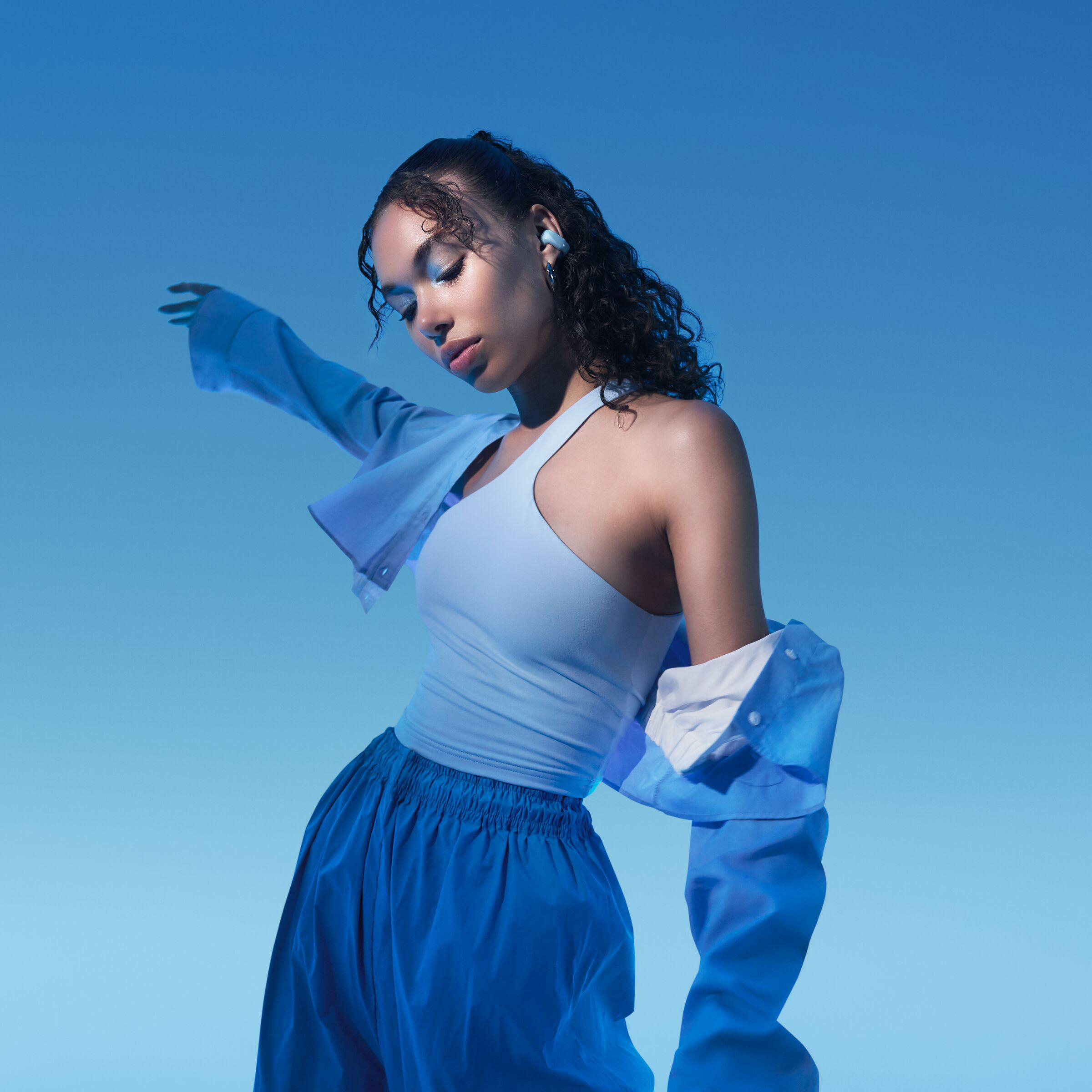 Eine Frau in blauem Outfit mit Ohrhörern posiert. Der Hintergrund ist einfarbig blau.
