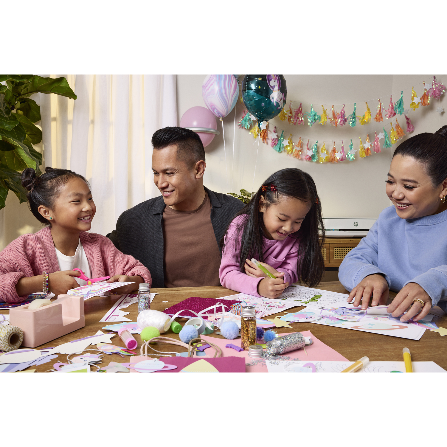 Famille à une table, bricolant. Ballons et décorations suspendus. Enfants souriants, parents. Fournitures diverses.