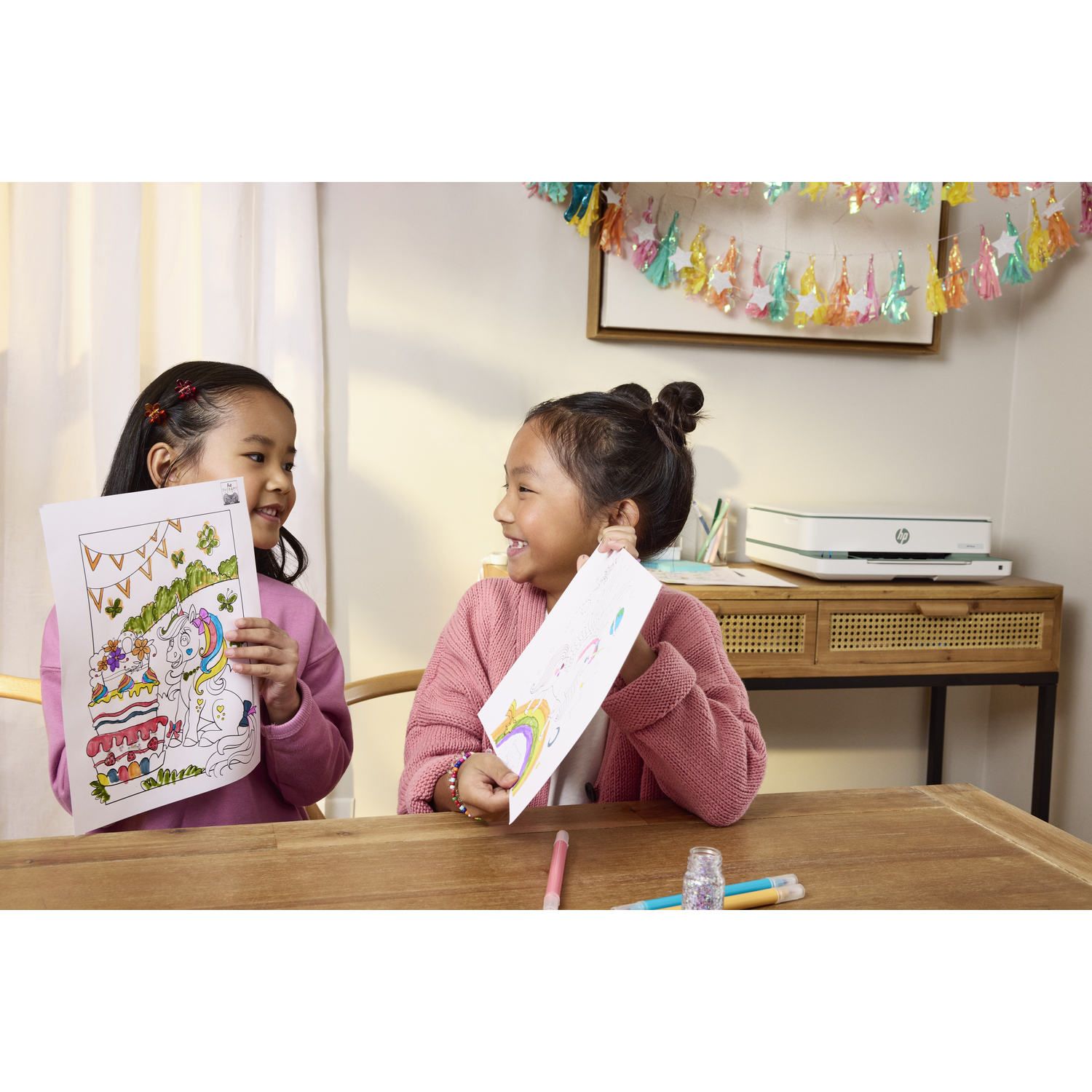 Deux jeunes filles souriantes tenant des dessins, avec une imprimante en arrière-plan.