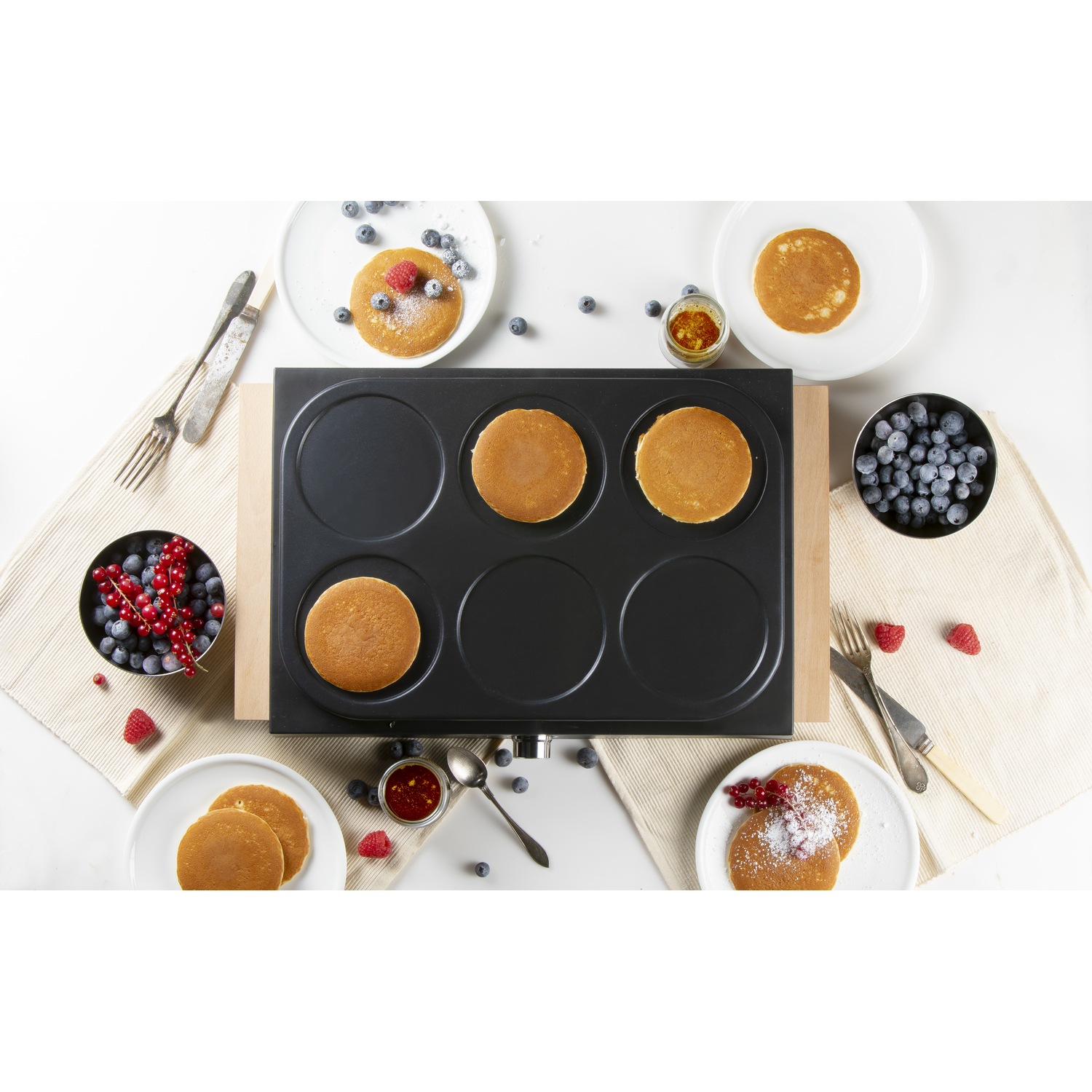Overzichtsfoto van een bakplaat voor zes pannenkoeken met pannenkoeken, fruit en kruiden op een witte tafel.
