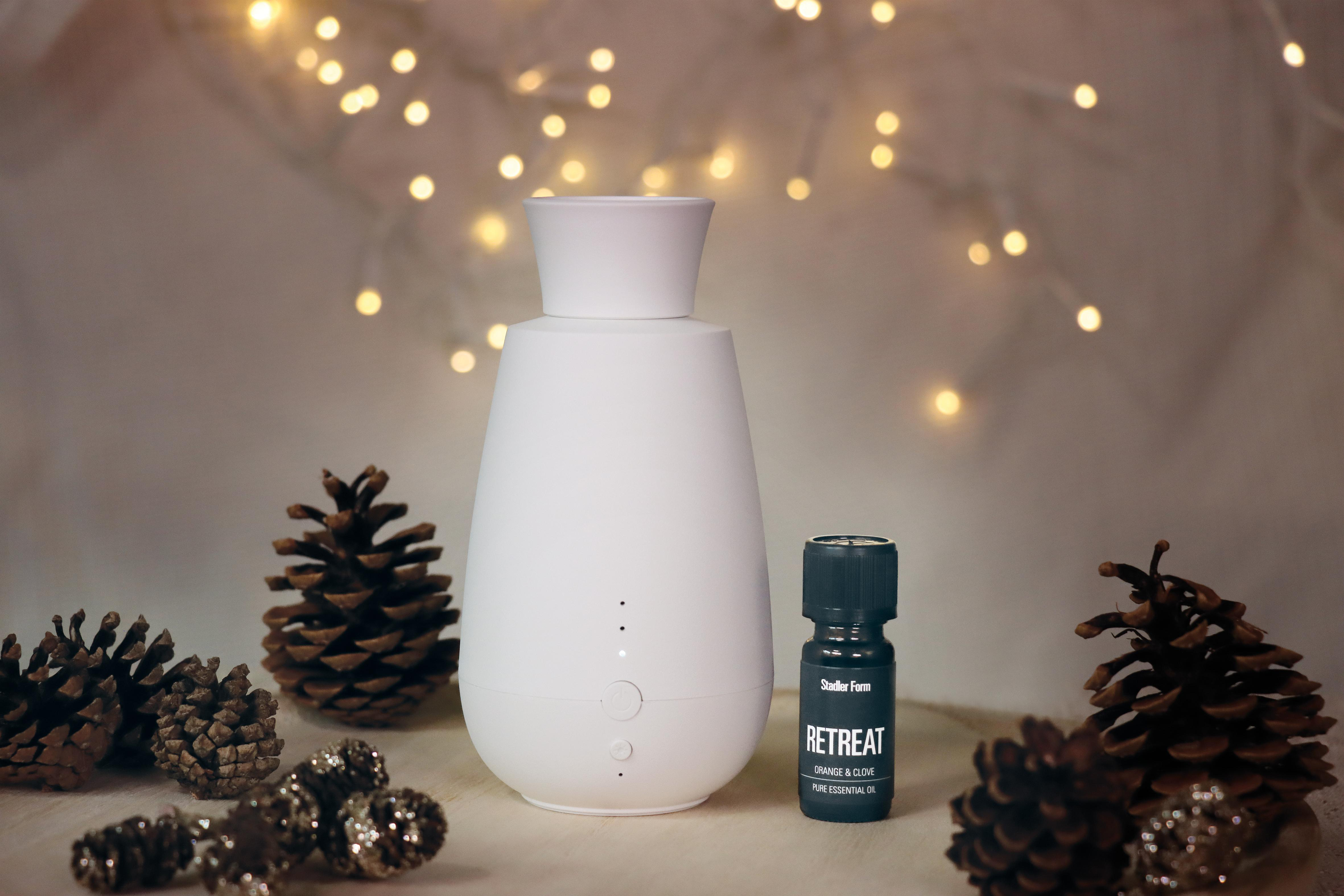 Weißer Diffusor für ätherische Öle mit 'Retreat'-Ölflasche und Tannenzapfen auf Holzoberfläche.