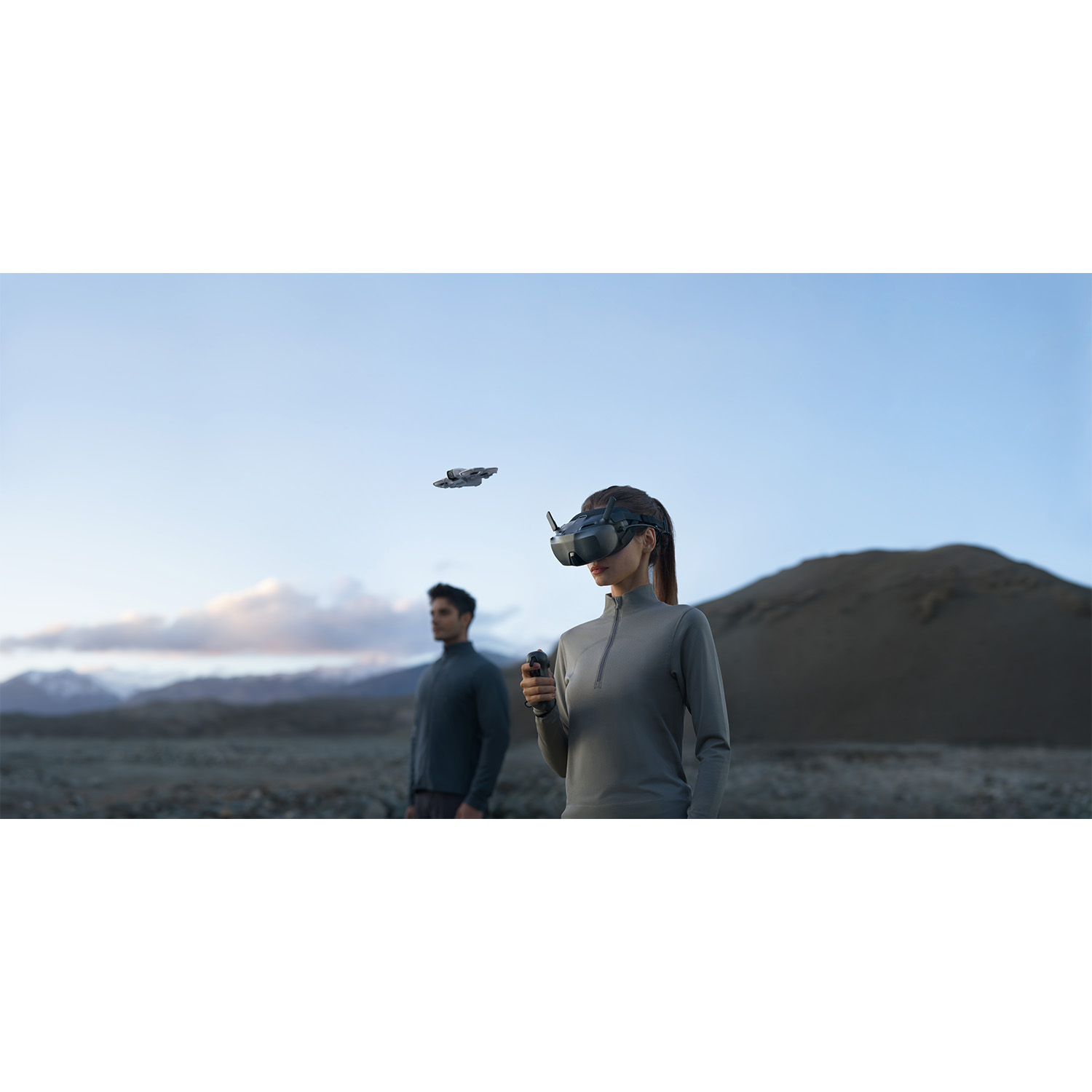 Donna con visore VR, controller, drone in volo. Uomo sullo sfondo. Cielo blu, scena montana.