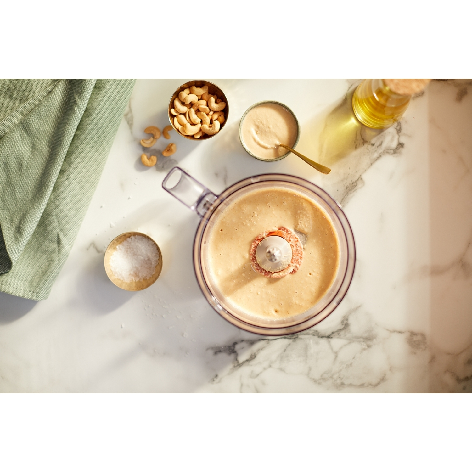 Robot da cucina con contenuto miscelato marrone chiaro. Anacardi e olio sono visibili nelle vicinanze, il tutto su marmo.