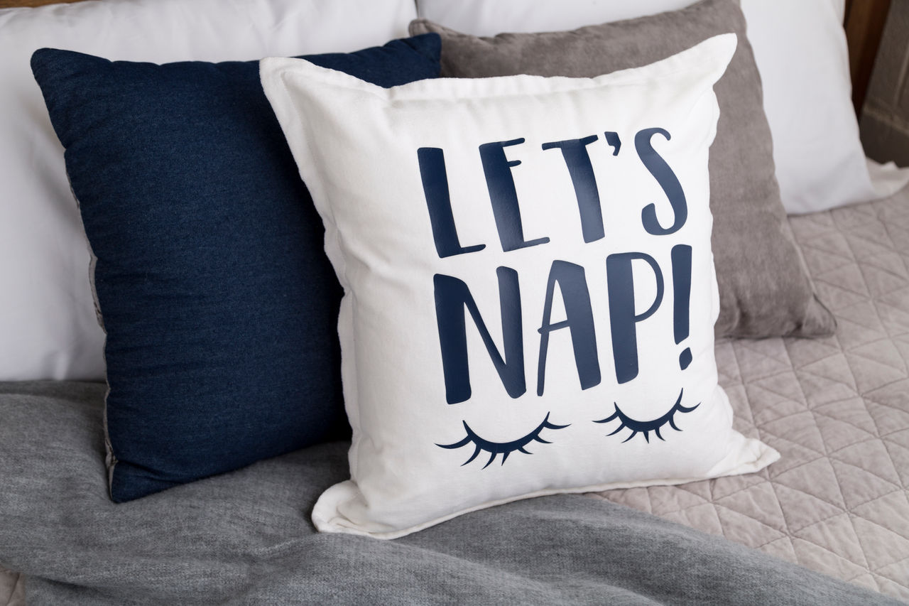 Weißes Kissen mit blauem Text 'LET'S NAP!' und Wimpern. Marine- und graue Kissen dahinter.