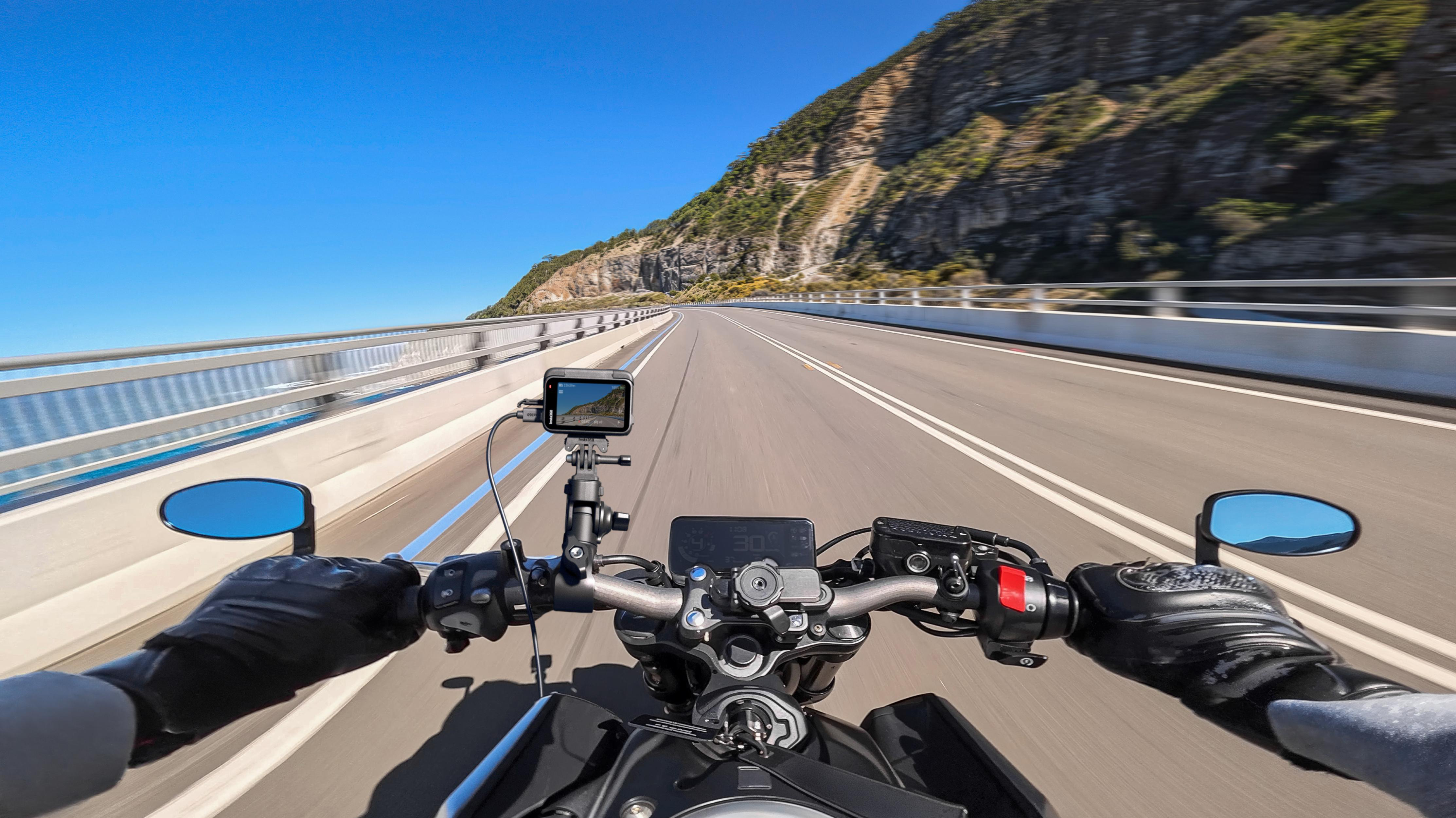 Motorrad-POV: Hände am Lenker, Straße, Bergkette und klarer blauer Himmel.