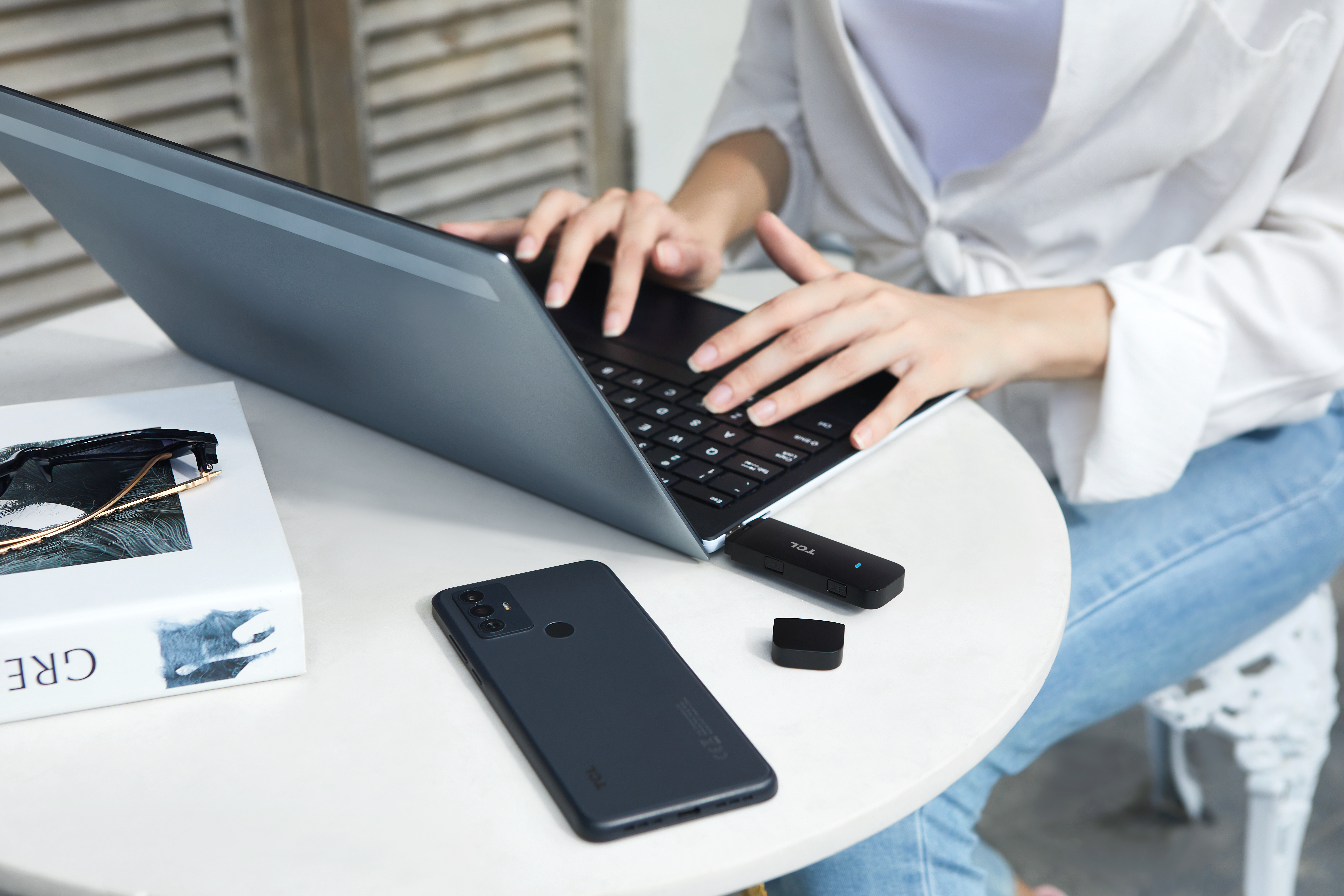 Osoba korzystająca z laptopa z telefonem, donglem i akcesoriami na białym stole.