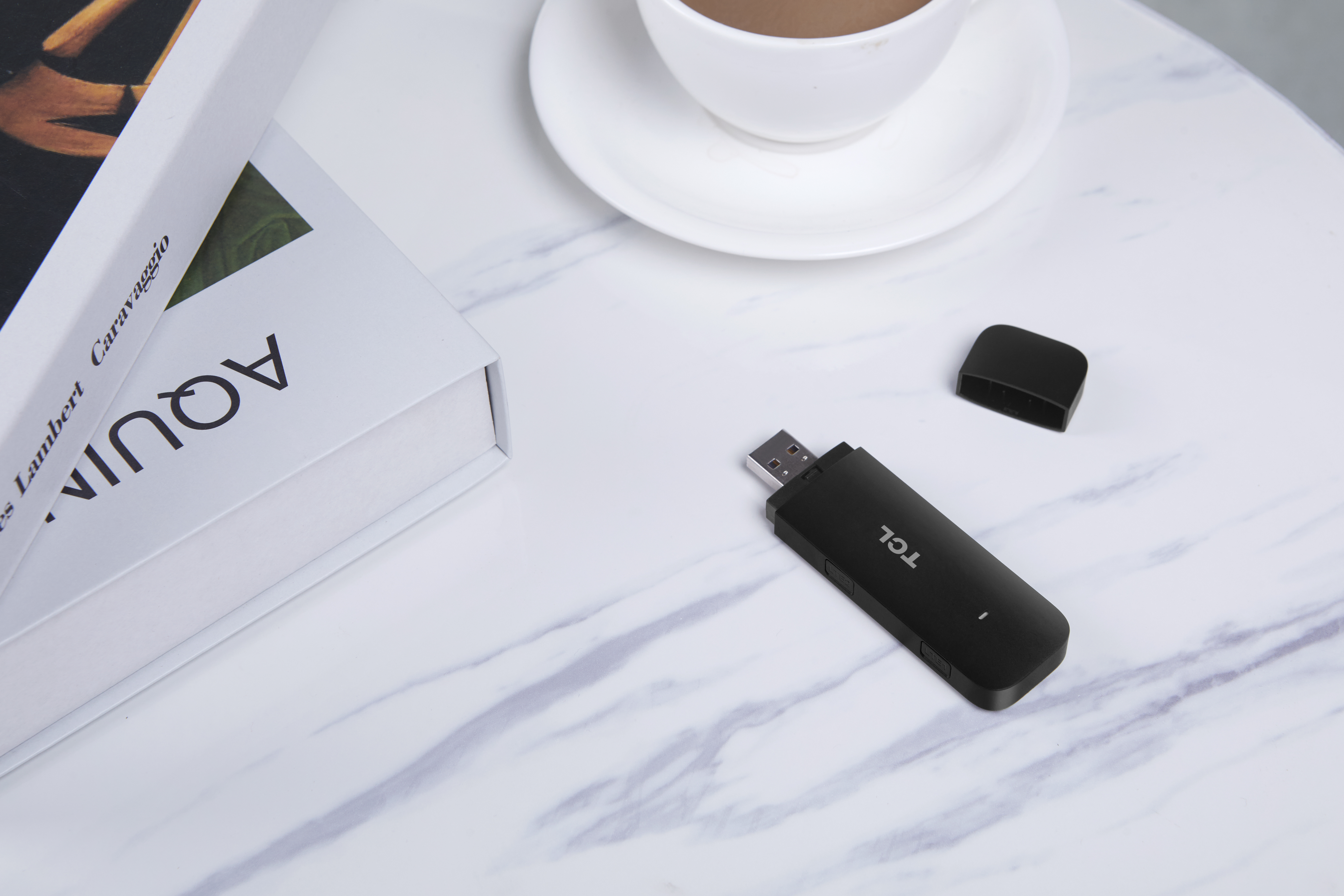 Czarny modem USB TCL i otwarta nasadka na białym marmurowym stole z książką i filiżanką kawy.