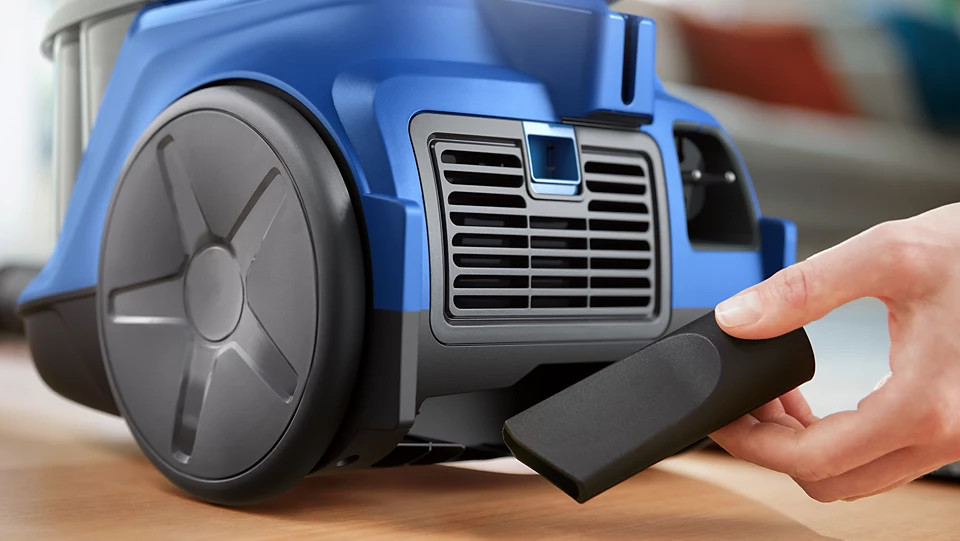 Niebieski odkurzacz z ręką sięgającą po czarny osprzęt do czyszczenia.