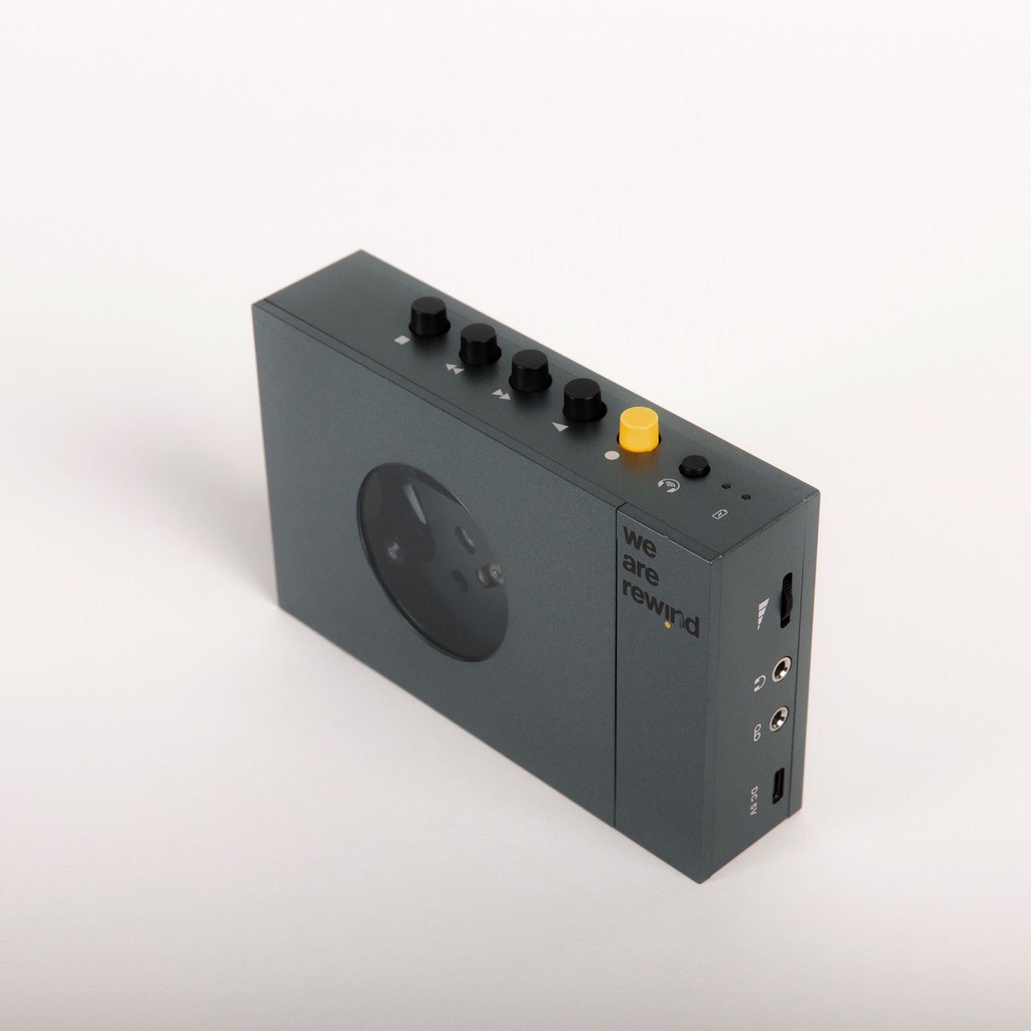 Lettore di cassette grigio scuro con pulsanti, un pulsante giallo e finestra rotonda, su sfondo bianco.