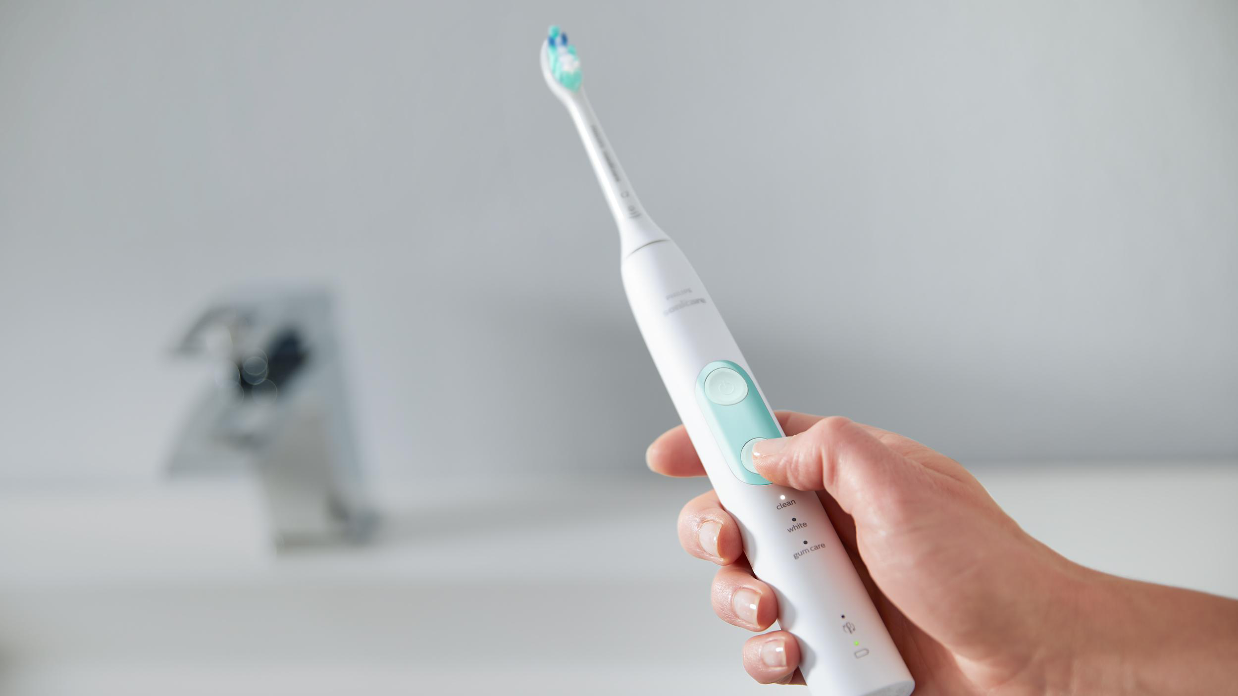 Hand hält eine weiße elektrische Zahnbürste. Wasser und Waschbecken im Hintergrund. Die Bürste ist an.
