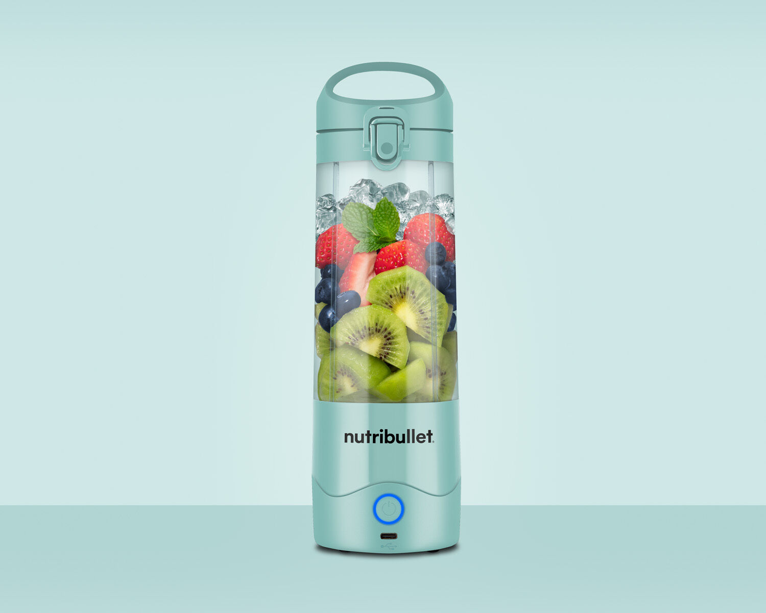 Una licuadora Nutribullet verde llena de hielo, fresas, arándanos, kiwis y hojas de menta.