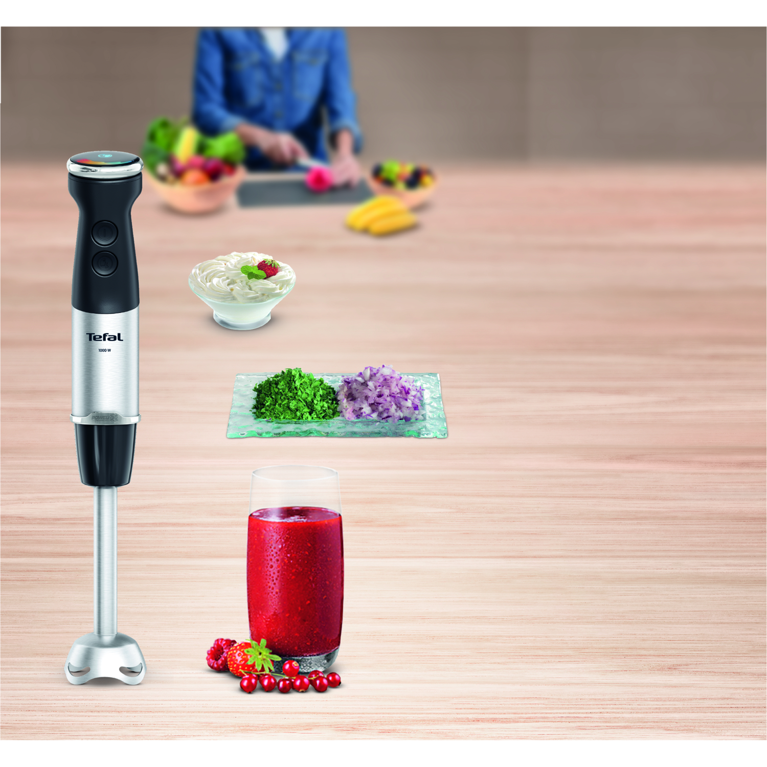 Staafmixer met ingrediënten, smoothie in glas, wazige persoon op achtergrond.