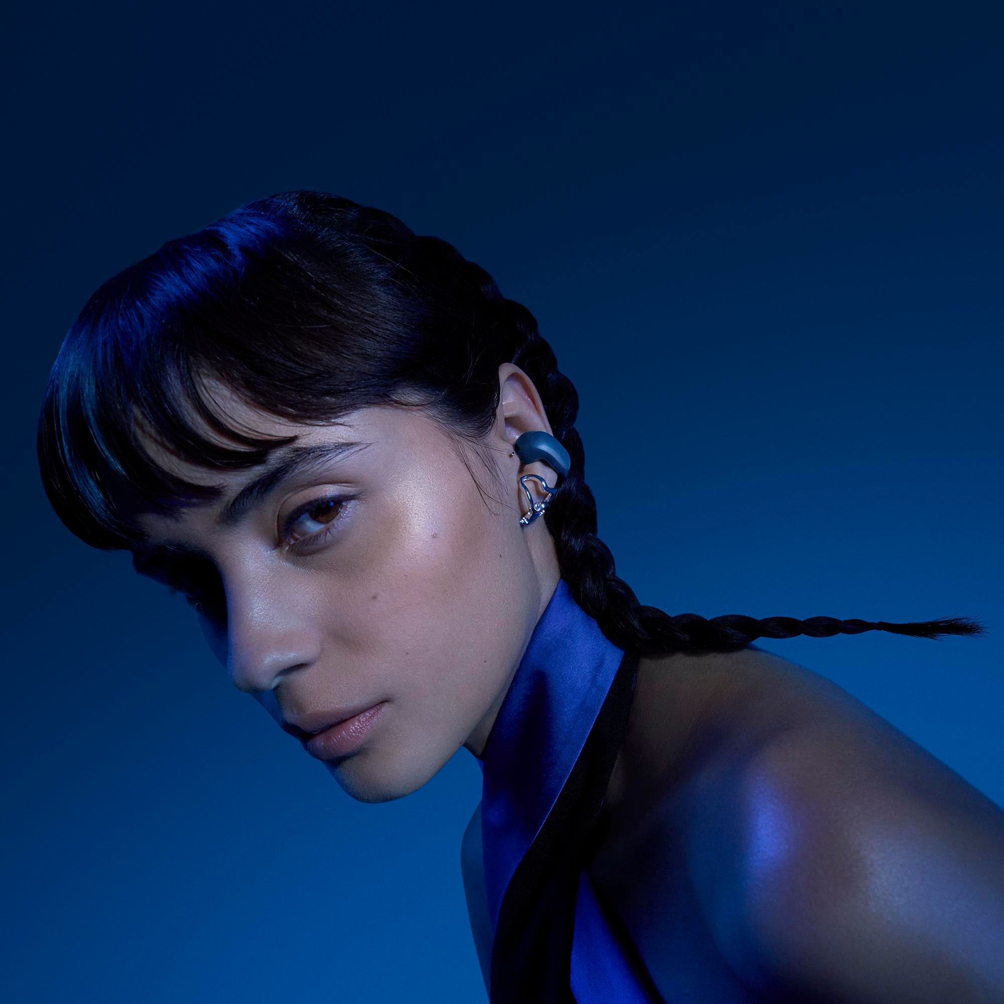 Eine Frau mit geflochtenen Haaren und auffälligem Schmuck am Ohr, vor blauem Hintergrund.