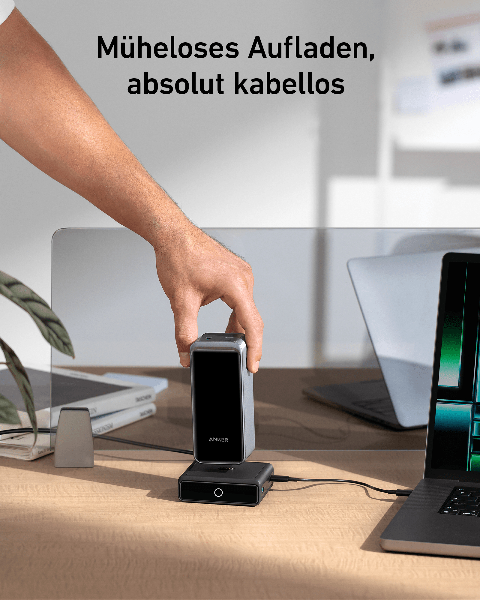 Eine Hand hält ein silbernes Ladegerät. Ein Laptop und ein weiteres Gerät im Hintergrund.