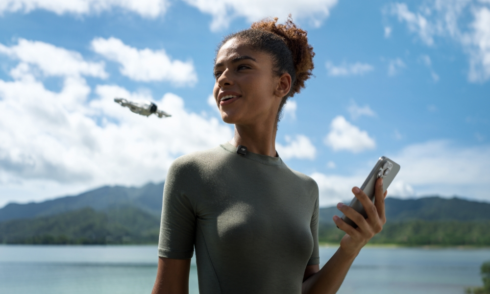 Uśmiechnięta kobieta trzymająca telefon, z latającym obiektem i jeziorem w tle.