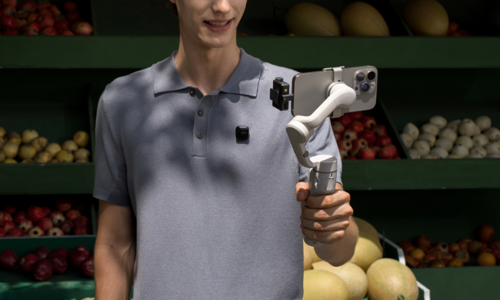 Mężczyzna trzymający smartfona przymocowanego do gimbala. Uśmiecha się przed zielonym sklepem.