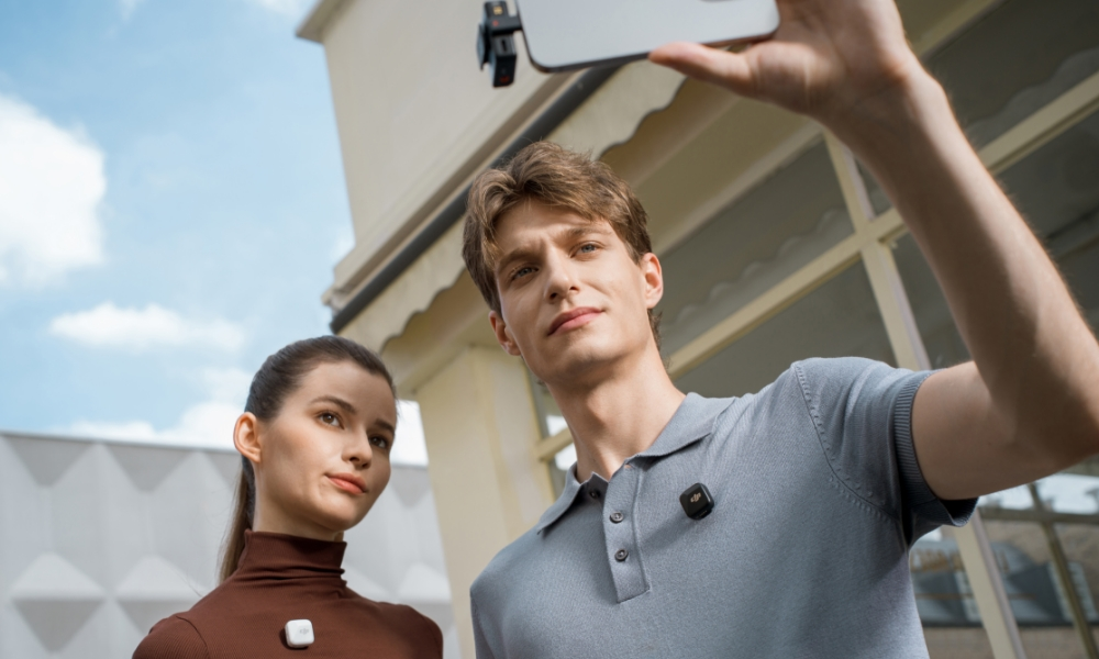 Dwie osoby, jedna trzyma telefon z mikrofonem. Tło to słoneczny dzień.