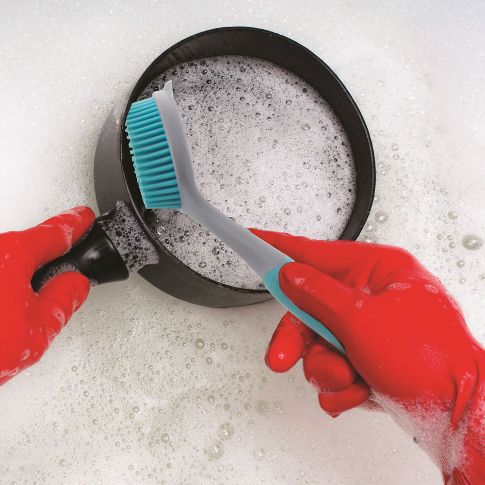 Manos con guantes rojos lavando una sartén con cepillo turquesa en agua jabonosa.