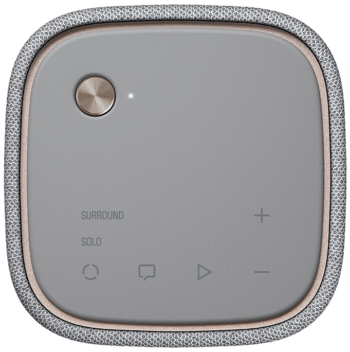 Szary głośnik audio. Posiada przycisk i regulację głośności. Otoczony szarą tkaniną.