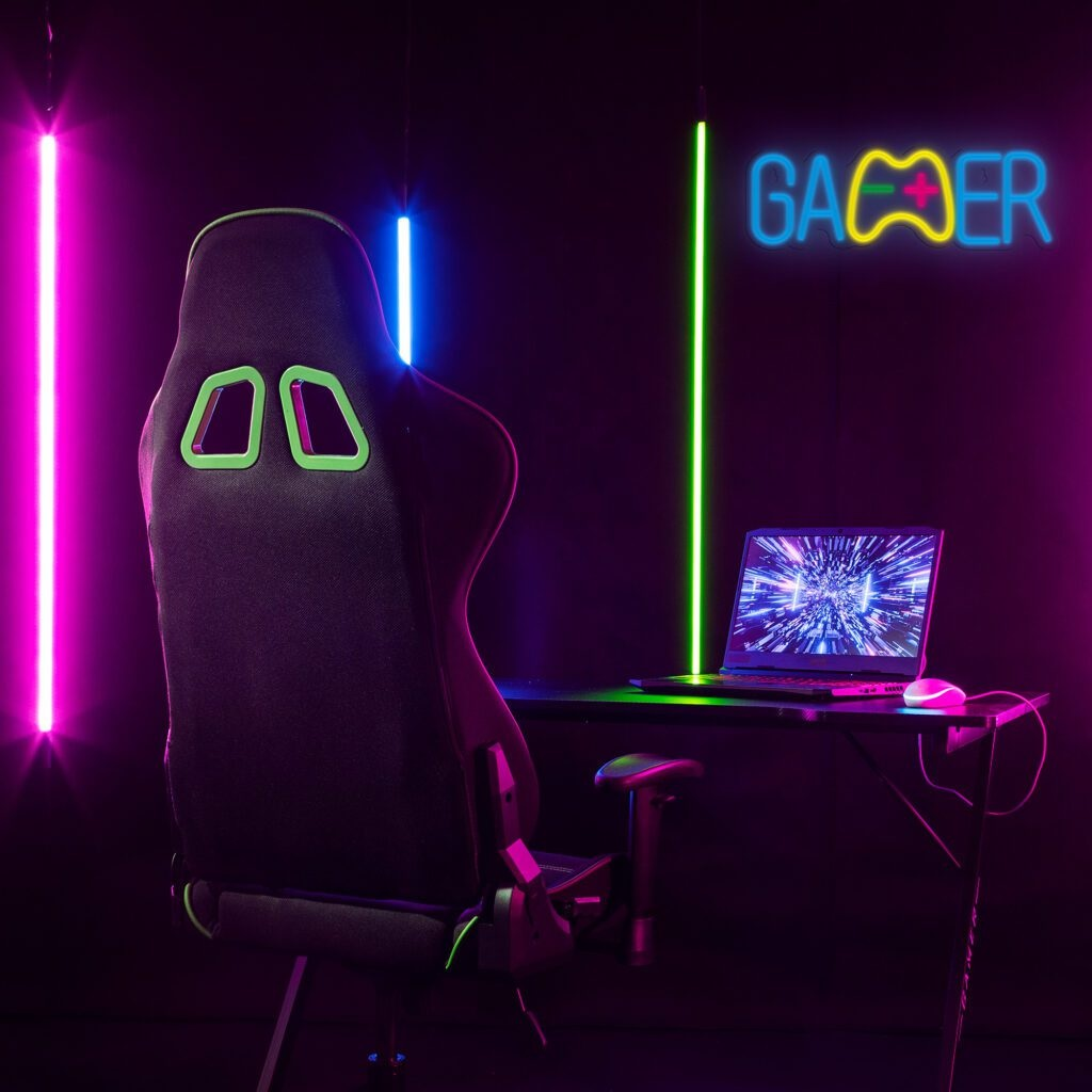 Konfiguracja gracza: czarny fotel, biurko z laptopem, neonowy znak 'GAMER' z ikoną kontrolera, różowe i niebieskie światła.