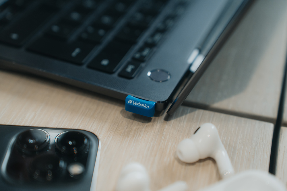 Bir dizüstü bilgisayara takılı mavi Verbatim USB sürücüsü. AirPods ve iPhone masada.