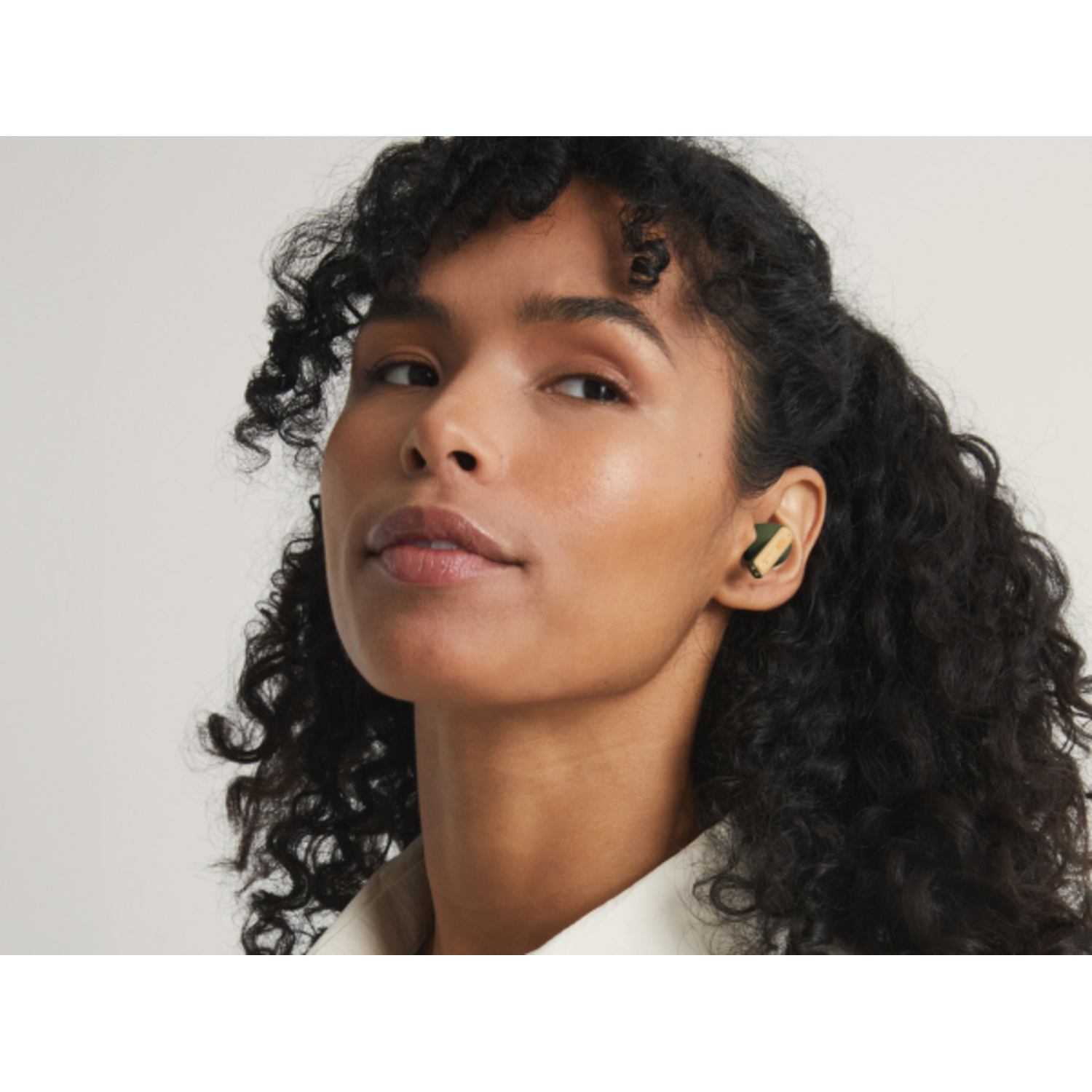 A woman with curly hair wears an earbud. The background is plain.