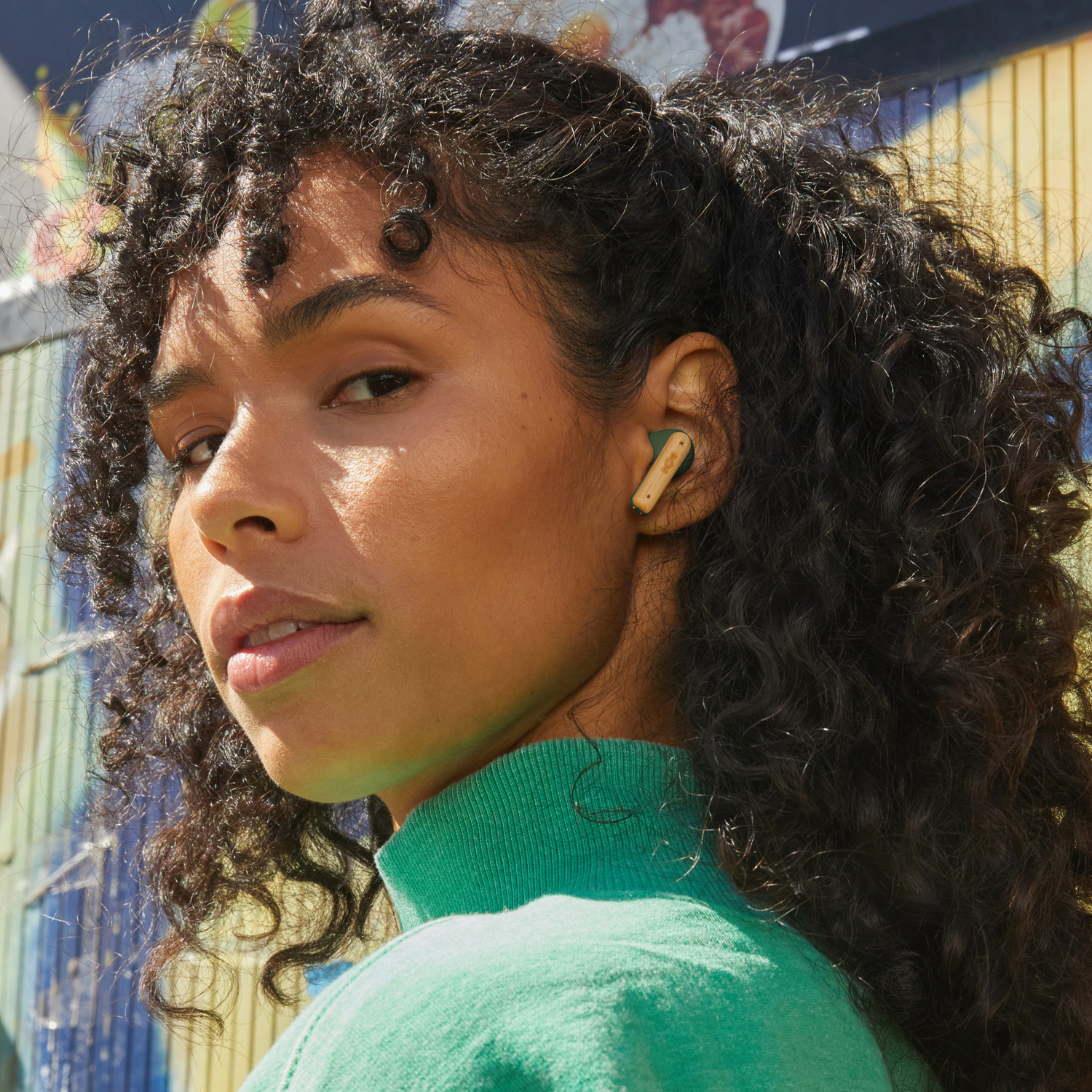 A woman with curly hair wearing green, has a earbud in her ear, looking at the camera.