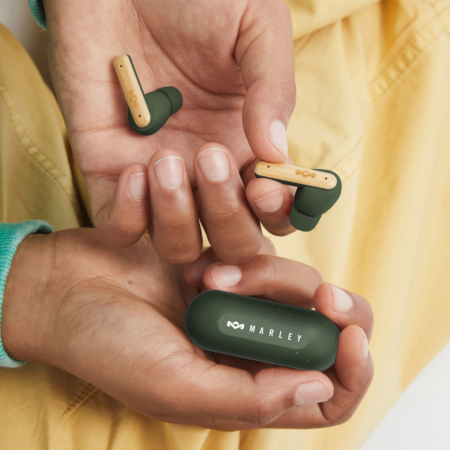 Hands holding wireless green earbuds and charging case, with a Marley logo on the case.