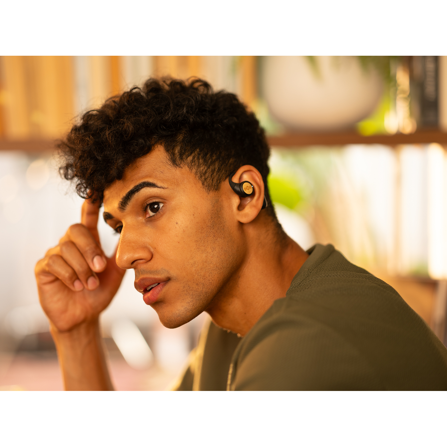 Un homme aux cheveux bouclés portant une chemise verte, avec un écouteur noir dans l'oreille droite. Visage concentré.