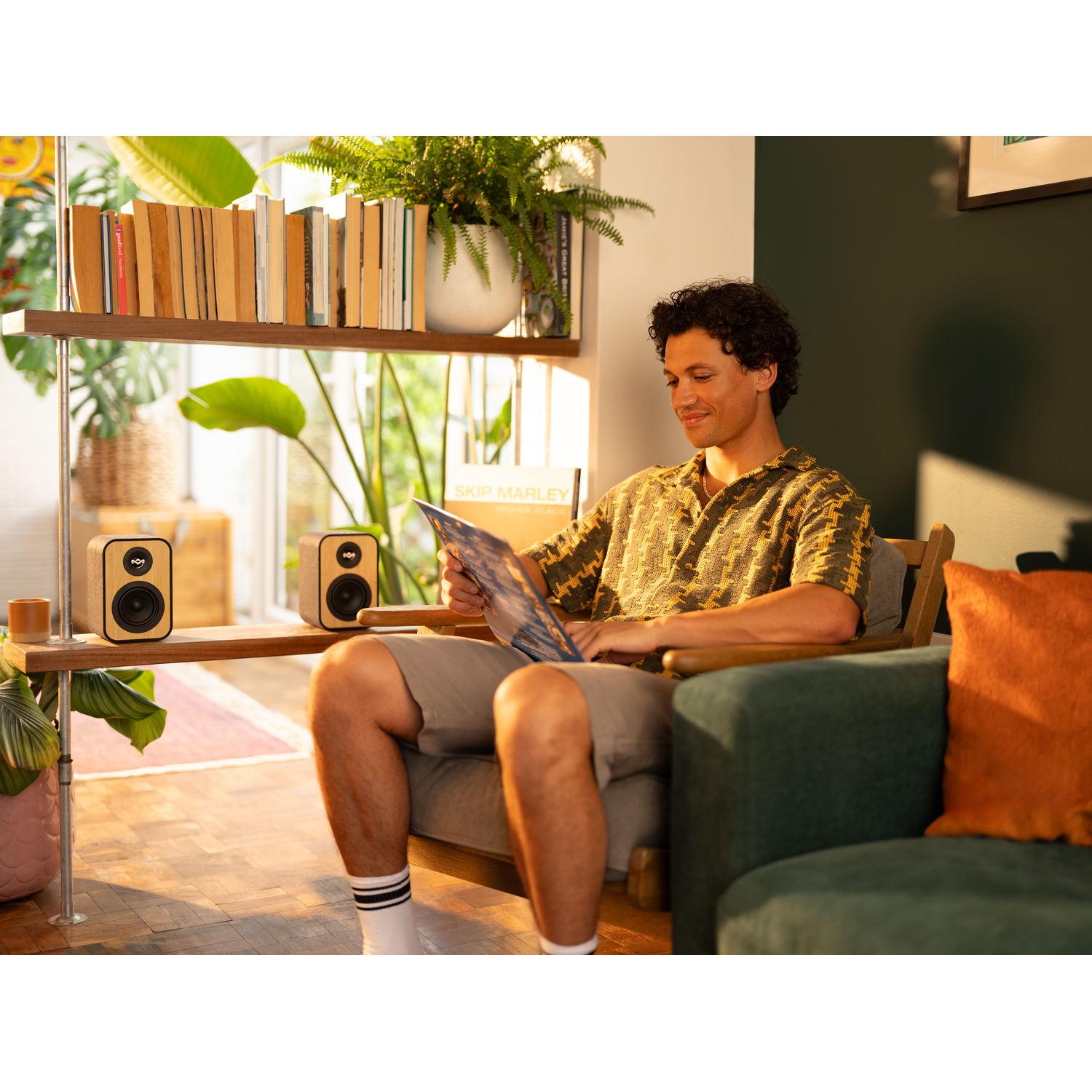 Man zit, houdt een plaat vast. Boekenplank met speakers, planten op de achtergrond.