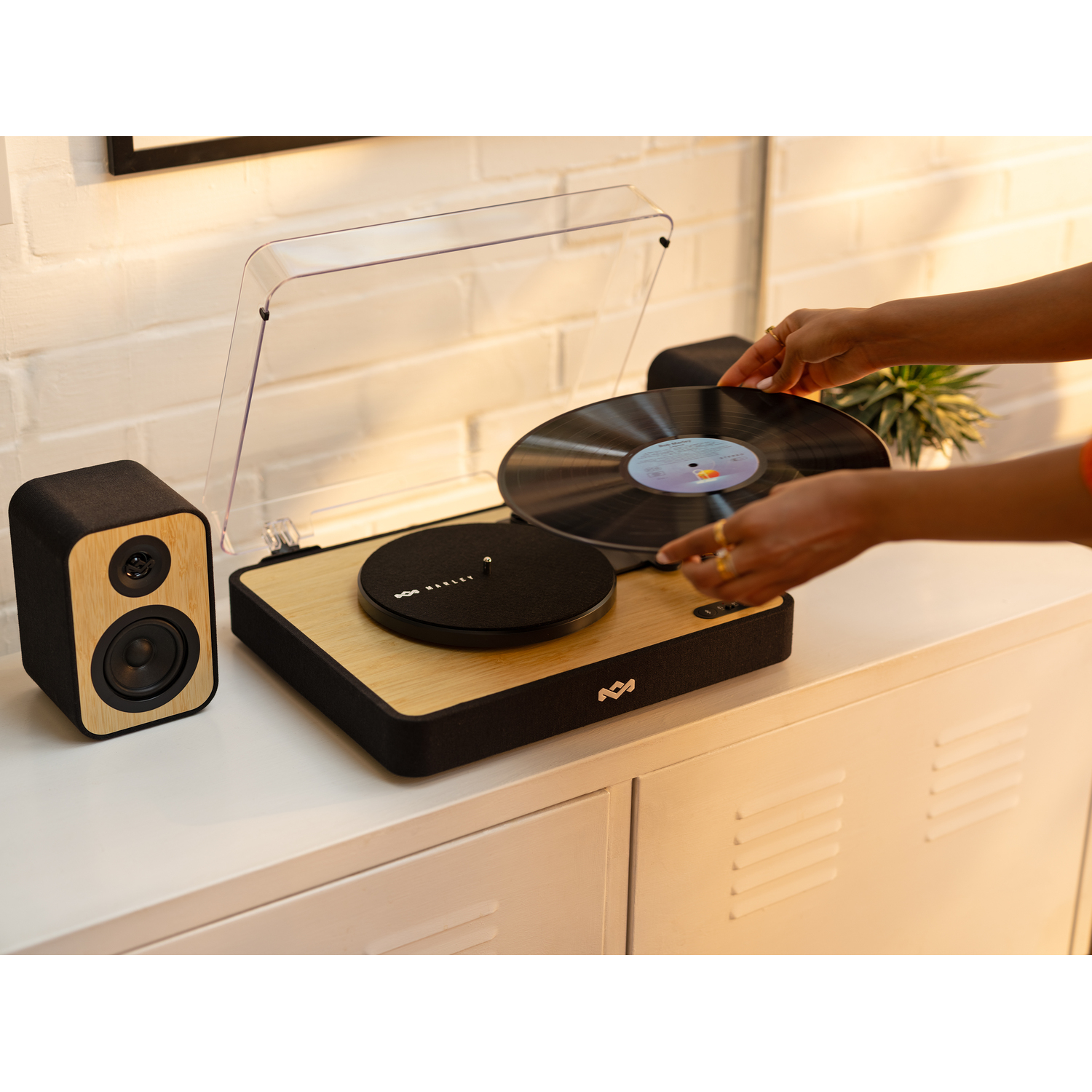 Een persoon die een vinylplaat op een houten platenspeler plaatst, naast een luidspreker. Witte kast op de achtergrond.