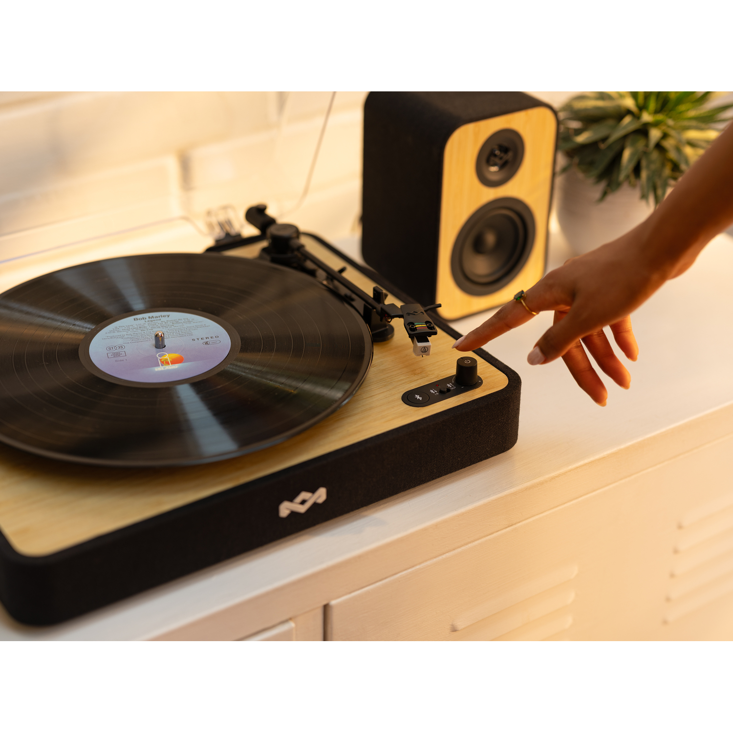 Een hand drukt op een knop op een houten platenspeler. Een luidspreker en een plant staan op de achtergrond. Een Bob Marley-plaat speelt.