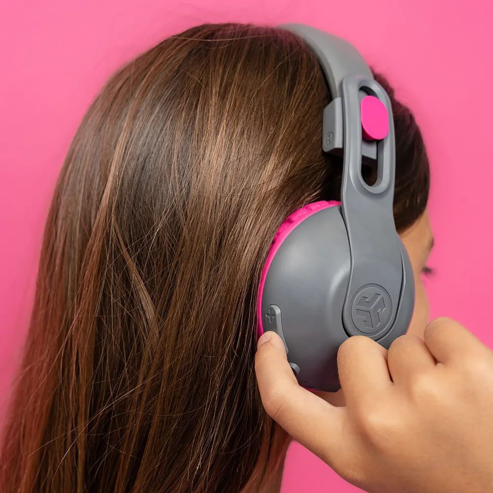 Ragazza con cuffie grigie e rosa, che tocca il pulsante del volume, sfondo rosa.