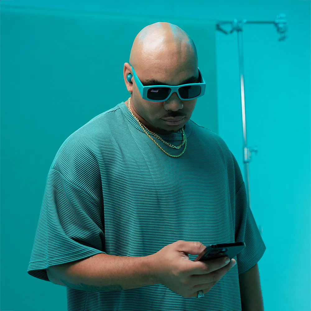 Mann mit rasiertem Kopf, Sonnenbrille und Hemd, hält ein Telefon. Türkisfarbener Hintergrund.