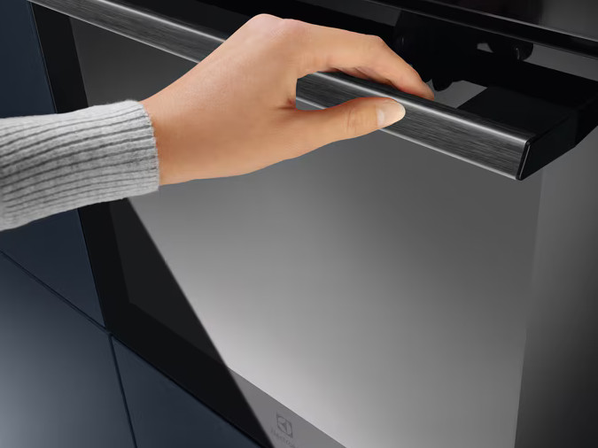 Ręka otwierająca drzwi piekarnika, z czarnym uchwytem i szklanymi drzwiami. Logo Electrolux widoczne.