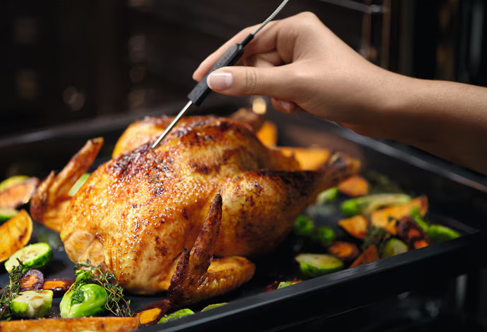 Dłoń wkładająca termometr do pieczonego kurczaka z warzywami.