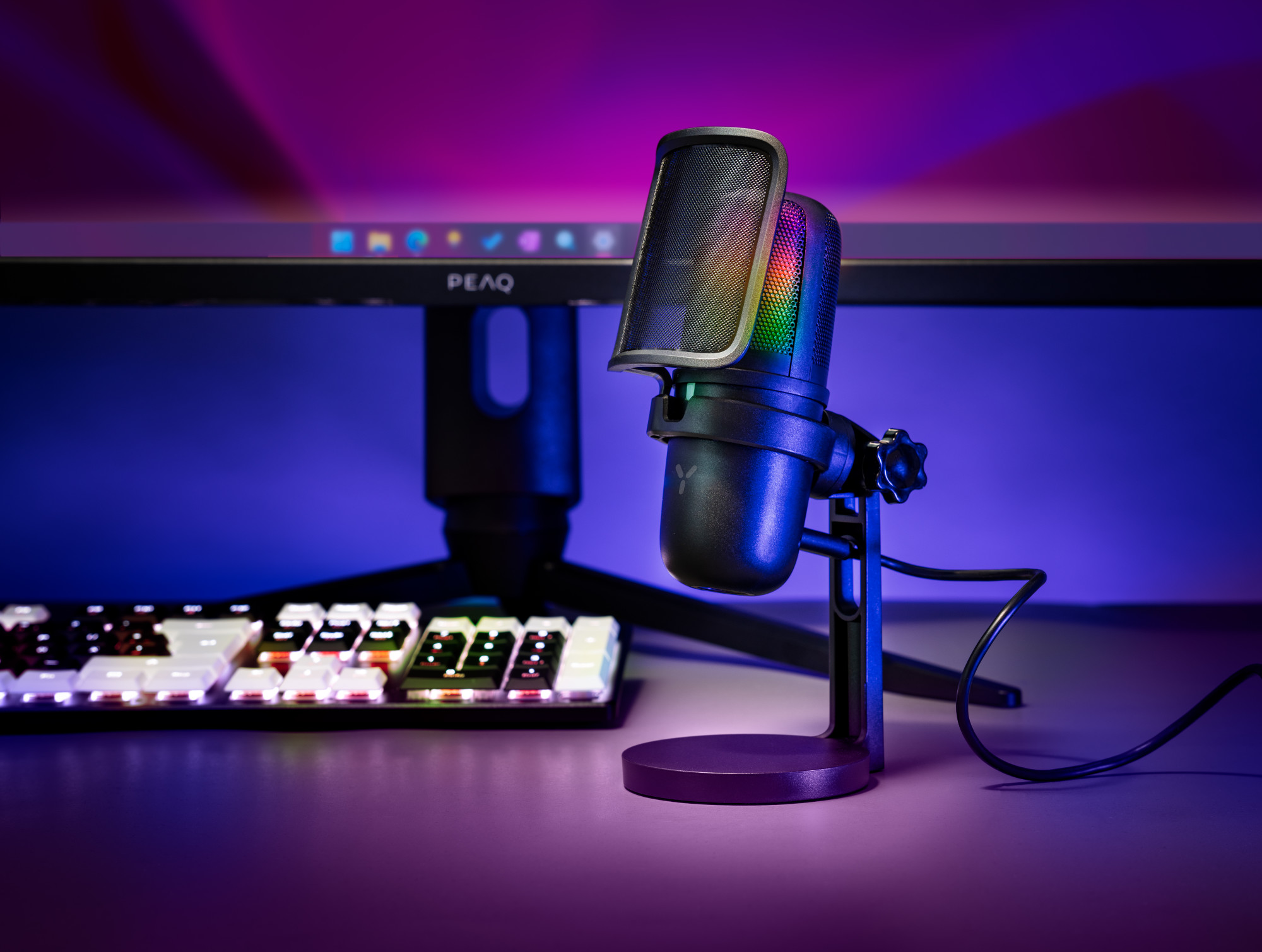 Czarny mikrofon ze stojakiem przed monitorem komputera i klawiaturą, oświetlony fioletowym i niebieskim światłem.