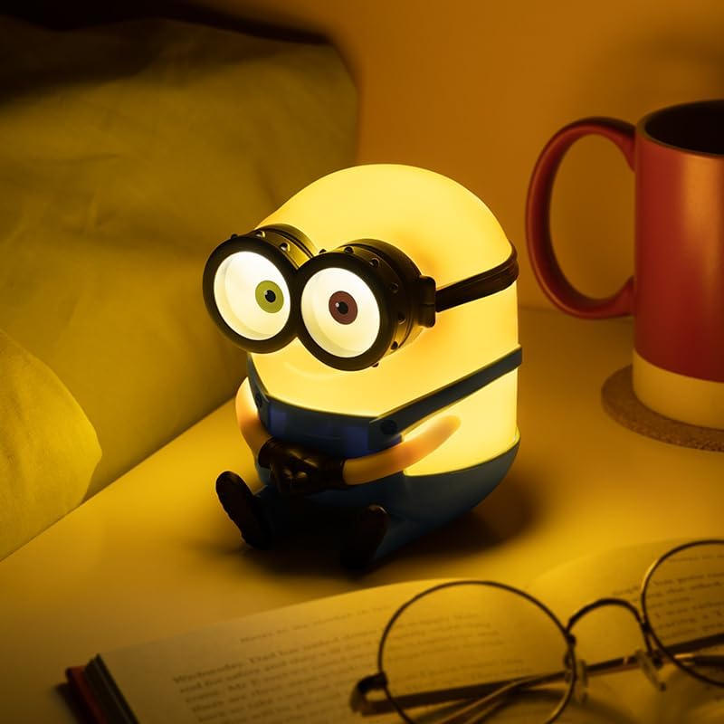 Egy világító Minion lámpa egy asztalon ül egy könyv, szemüveg és egy bögre mellett. A lámpának két szeme van, sárga és kék.