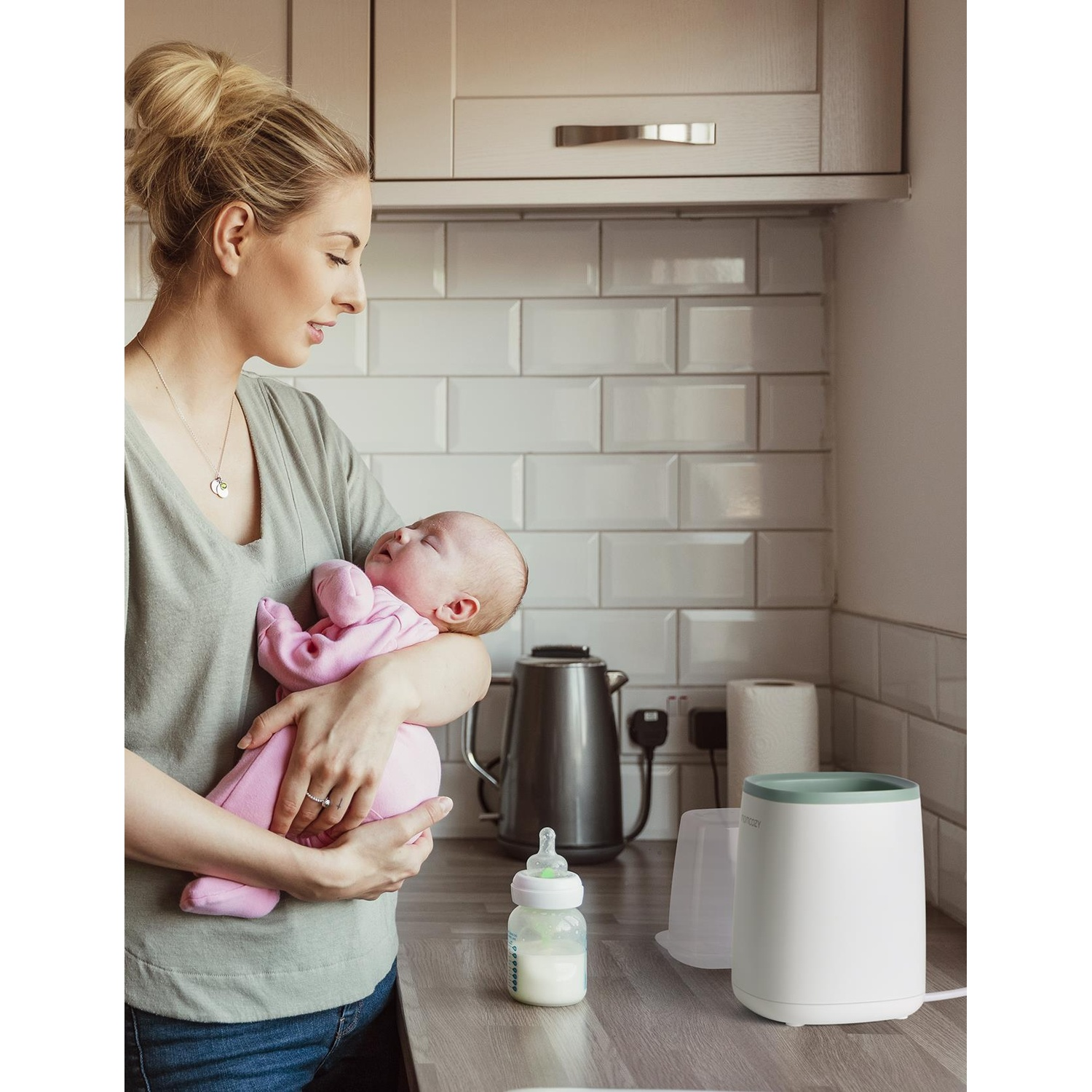 Una donna tiene in braccio un bambino vestito di rosa. Un biberon e uno scaldabiberon sono sul bancone. Cucina sullo sfondo.