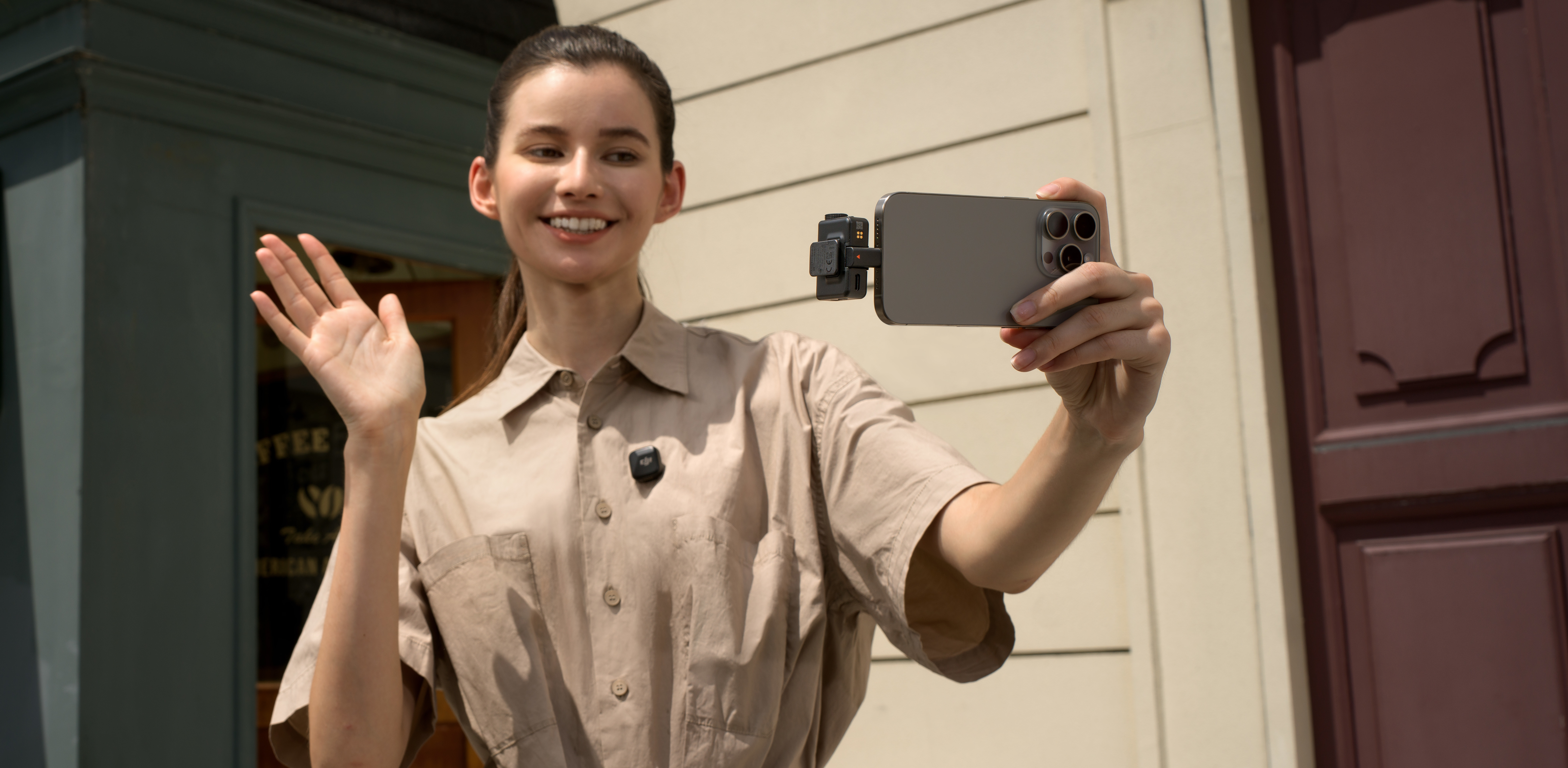 Femme salue, tenant un téléphone avec accessoire caméra. Chemise beige, bâtiment derrière.