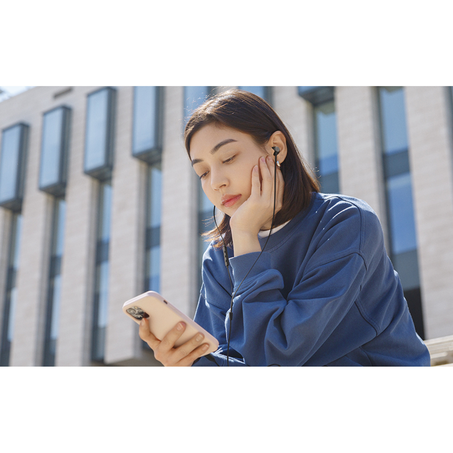 Vrouw kijkt naar telefoon, draagt oortjes, zit buiten.