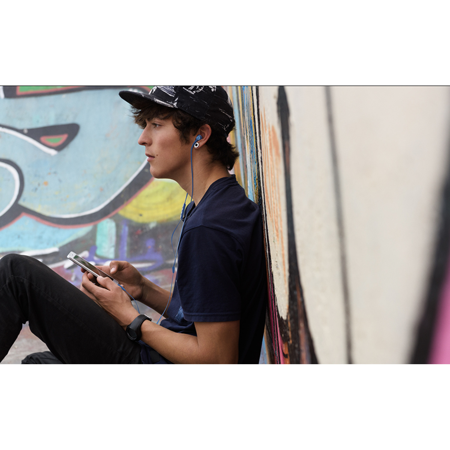 Een jonge man met oortelefoons en een telefoon zit bij een graffiti-muur.