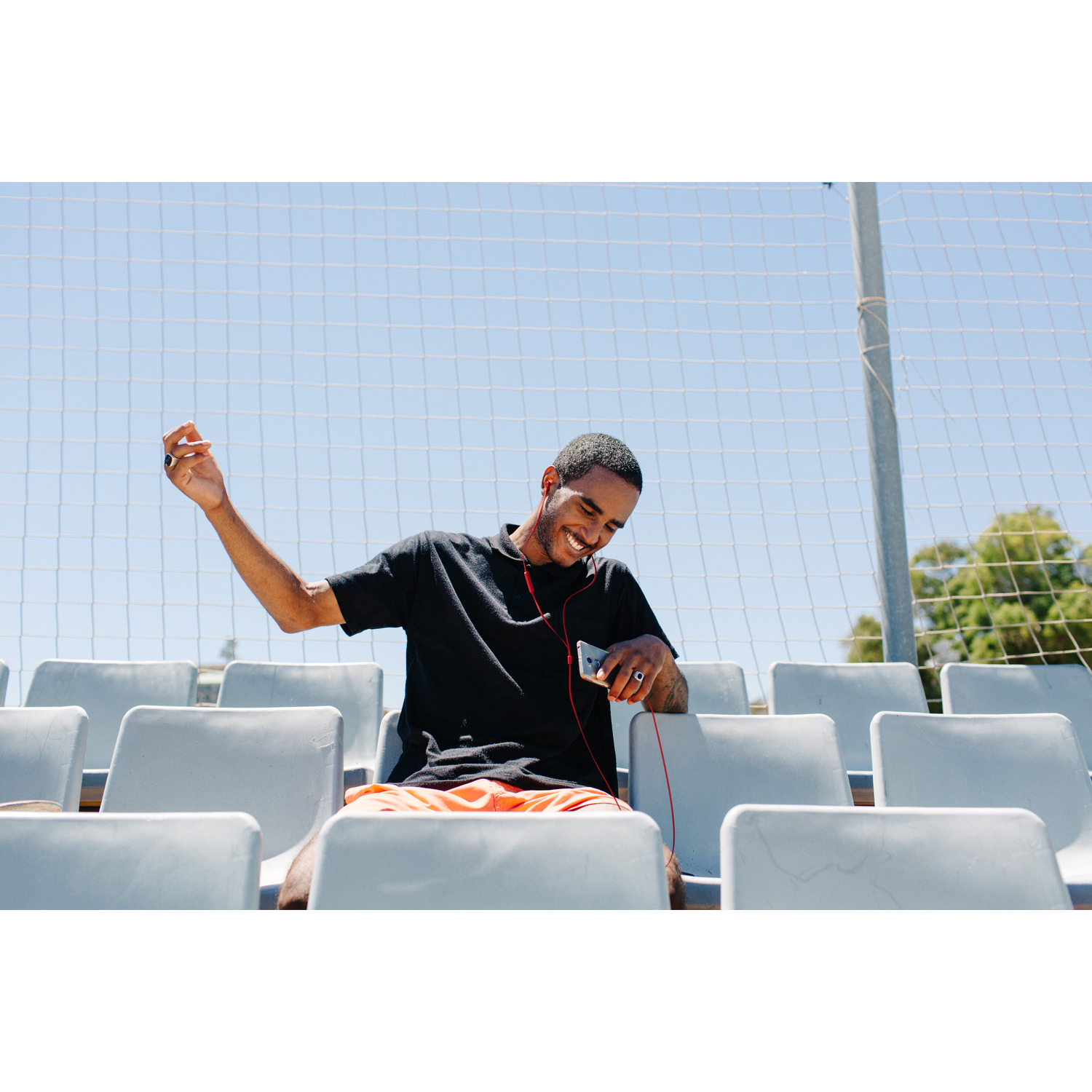 Een man met een zwart shirt en een oranje short glimlacht, luistert naar muziek op zijn telefoon in een stadion.