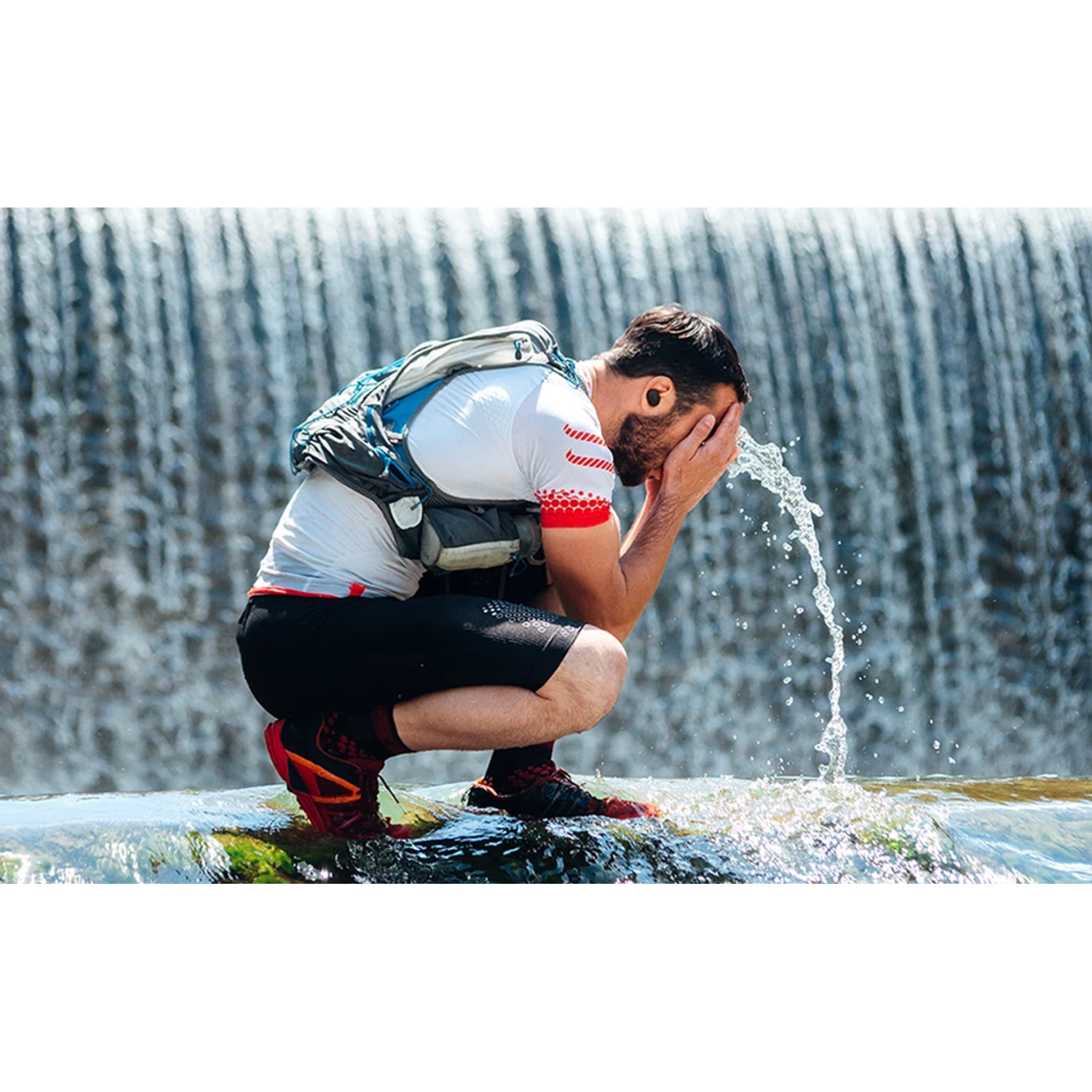 Man washing face with water from a waterfall. Black shorts, white shirt, blue backpack.