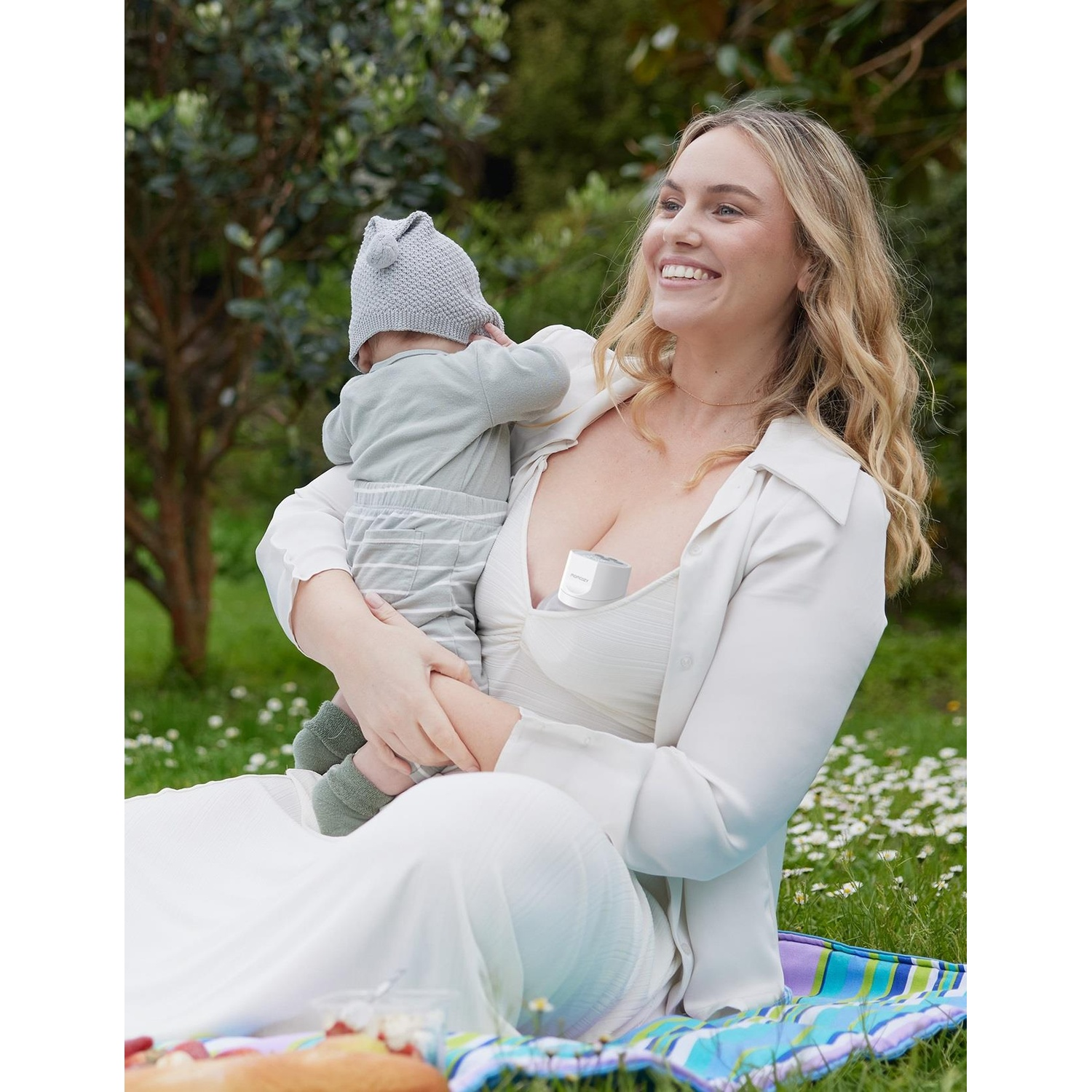 Donna che tiene bambino, camicia bianca, con tiralatte nel reggiseno, in un parco.