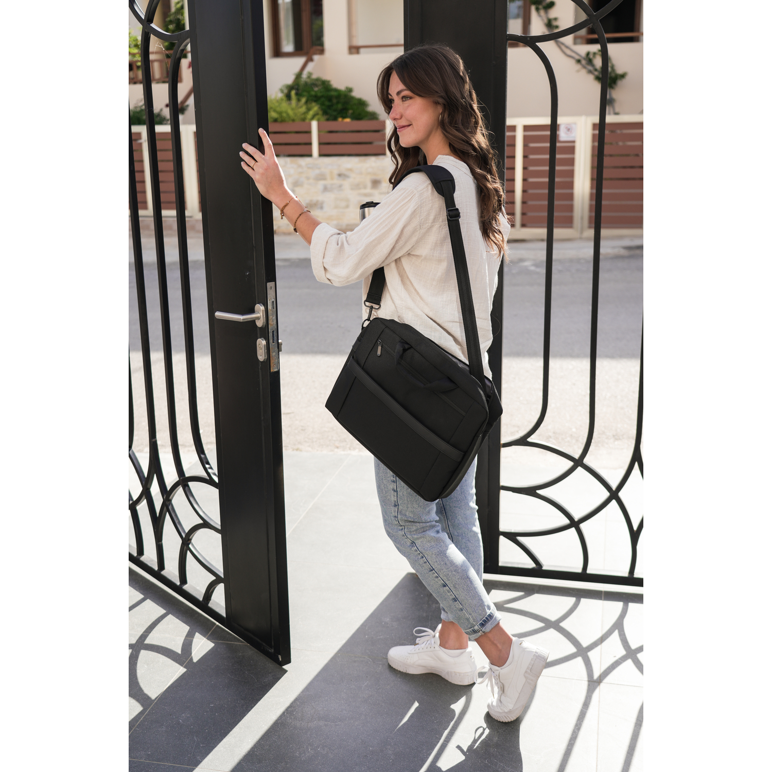 Woman in white shirt, jeans, white shoes opens black gate, black bag on shoulder.